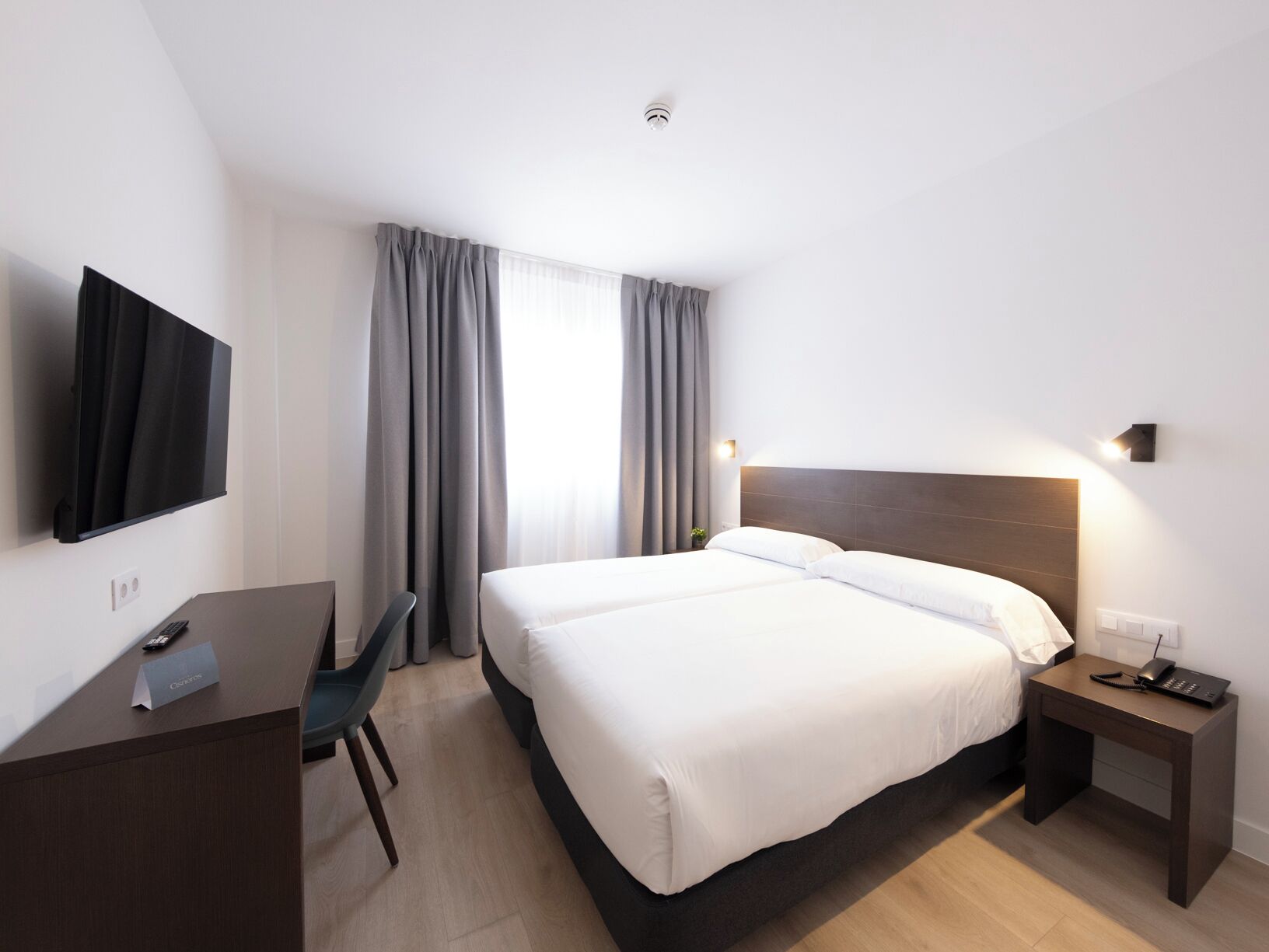 A hotel room with a bed, a desk, a chair, a TV, and a bedside table with a phone.