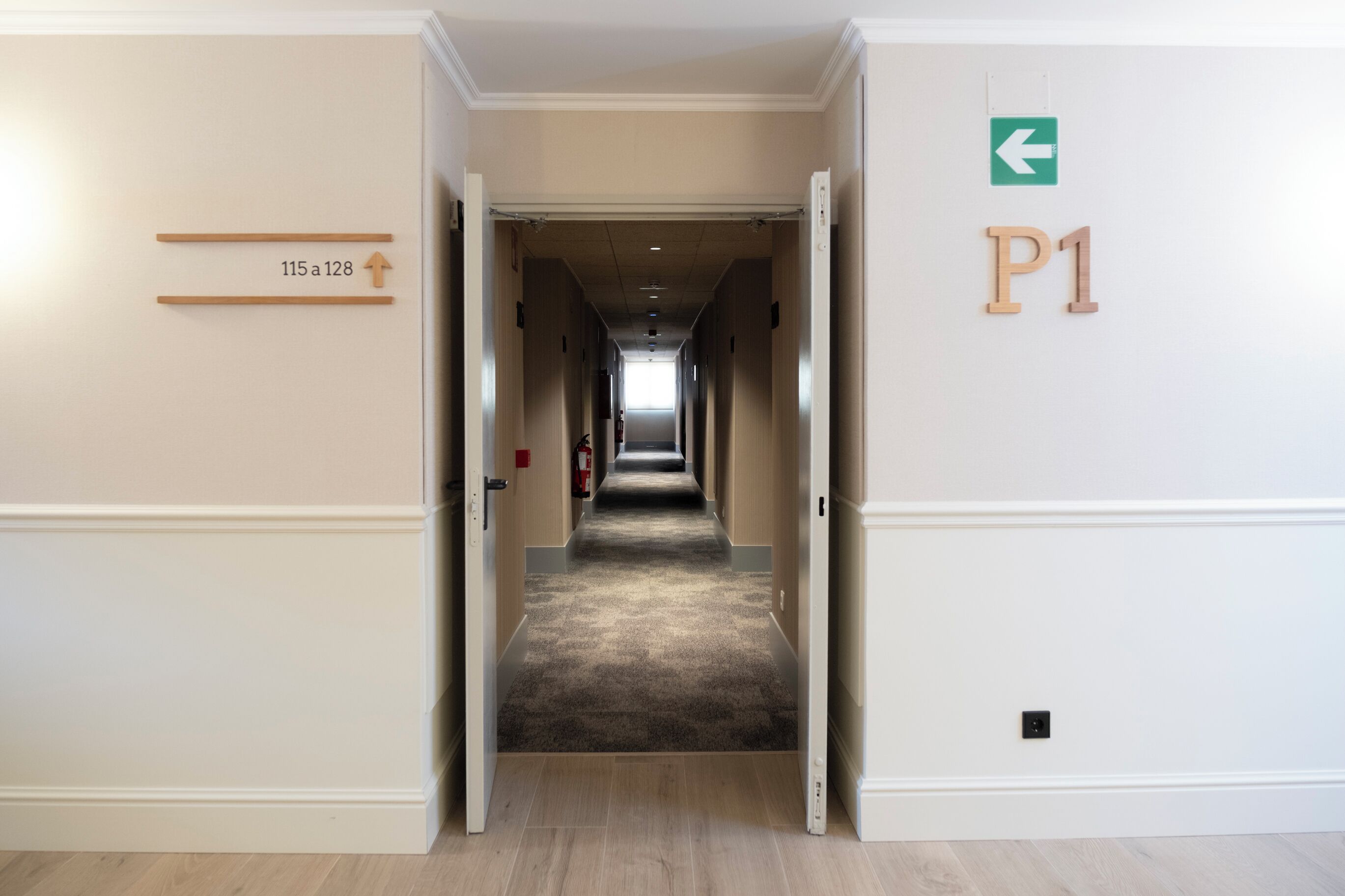 Een hotelgang met een vloerbedekking, een brandblusser en een bord dat de richting naar kamer P1 aangeeft.
