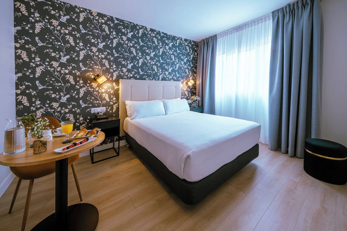 Une chambre d’hôtel avec un grand lit, une table en bois sur laquelle sont disposés des articles pour le petit-déjeuner, une lampe fixée au mur et une fenêtre avec des rideaux.