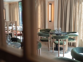 Interior de restaurante con mesas redondas, sillas tapizadas de verde y una pared con diseño.