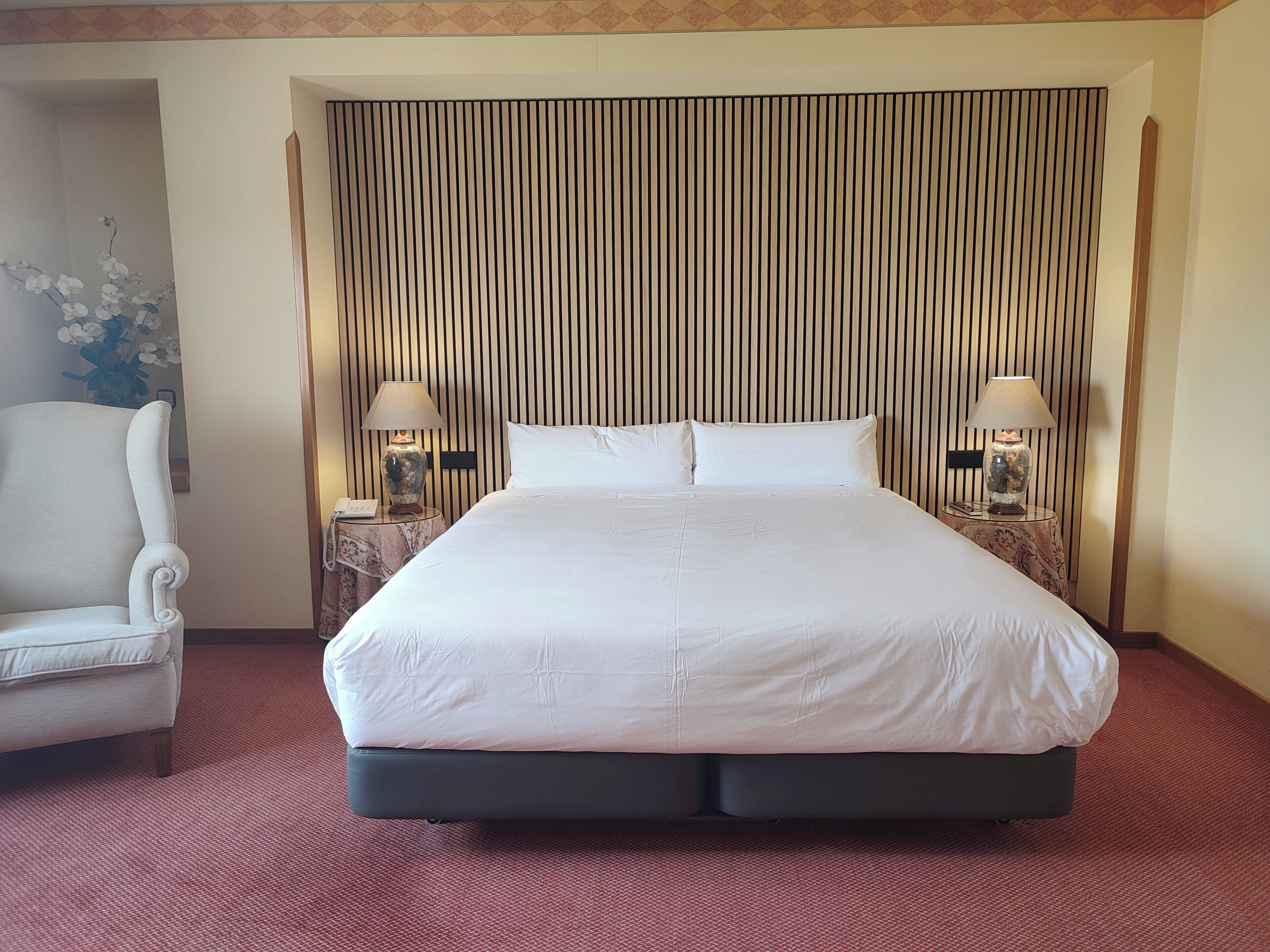 A hotel room with a large bed, two bedside tables with lamps, a chair, and a decorative wall panel.
