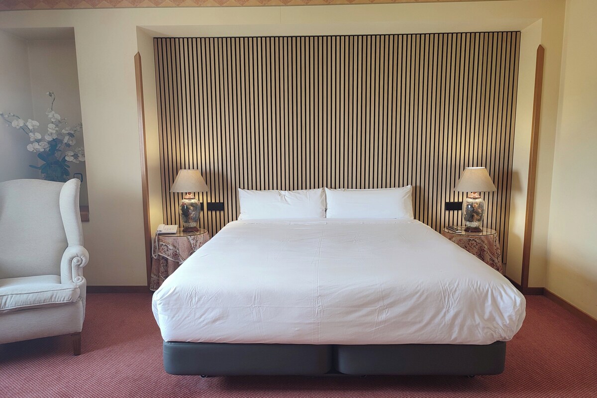 Habitación de hotel con una cama grande, dos mesitas de noche con lámparas, una silla y un panel decorativo en la pared.