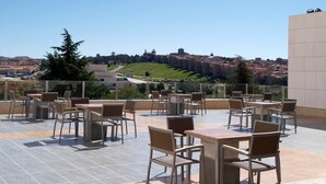 A rooftop terrace with outdoor seating, a clear view of a cityscape, and a large building in the background. - Hotel Sercotel Cuatro Postes (Ávila)
