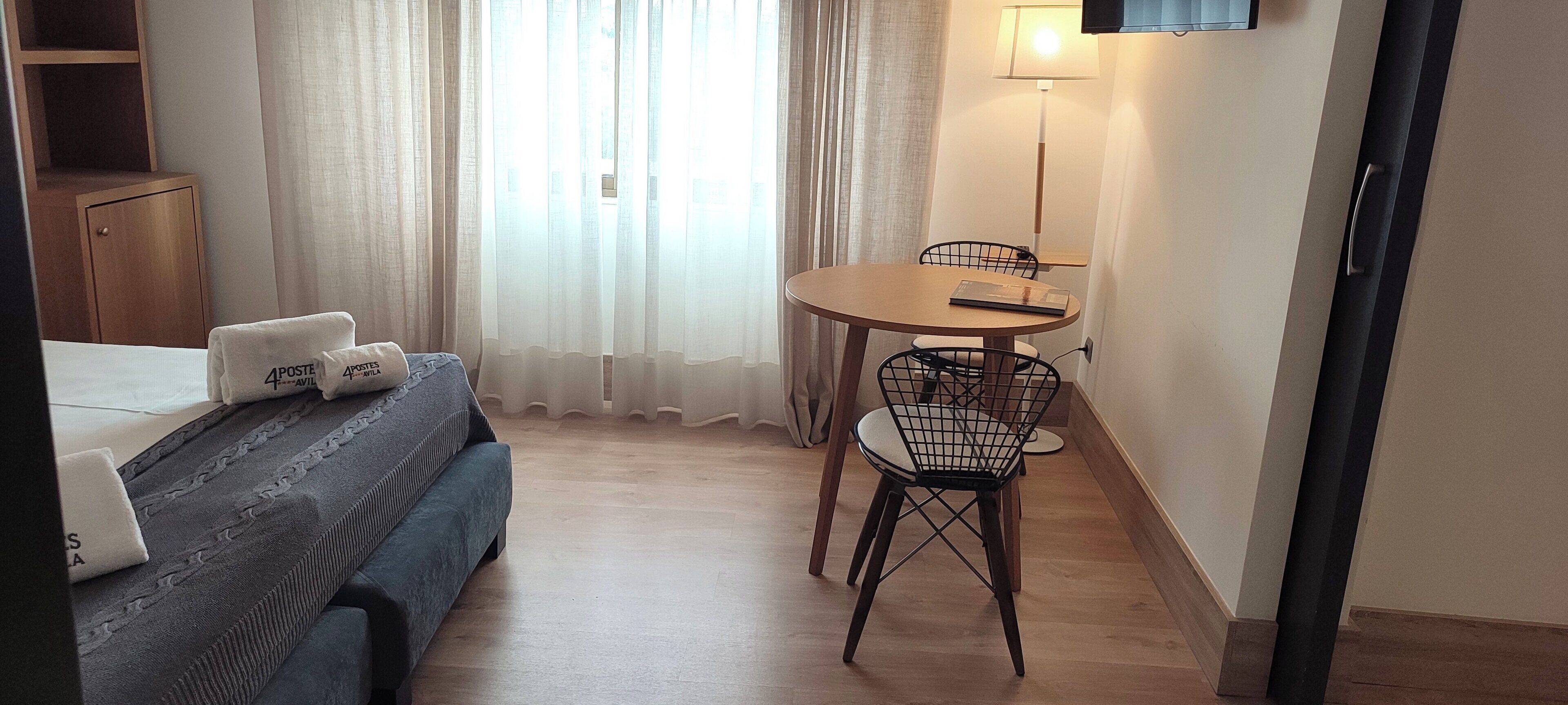 Een hotelkamer met een bed, een klein tafeltje met een stoel, een lamp en een raam met gordijnen.
