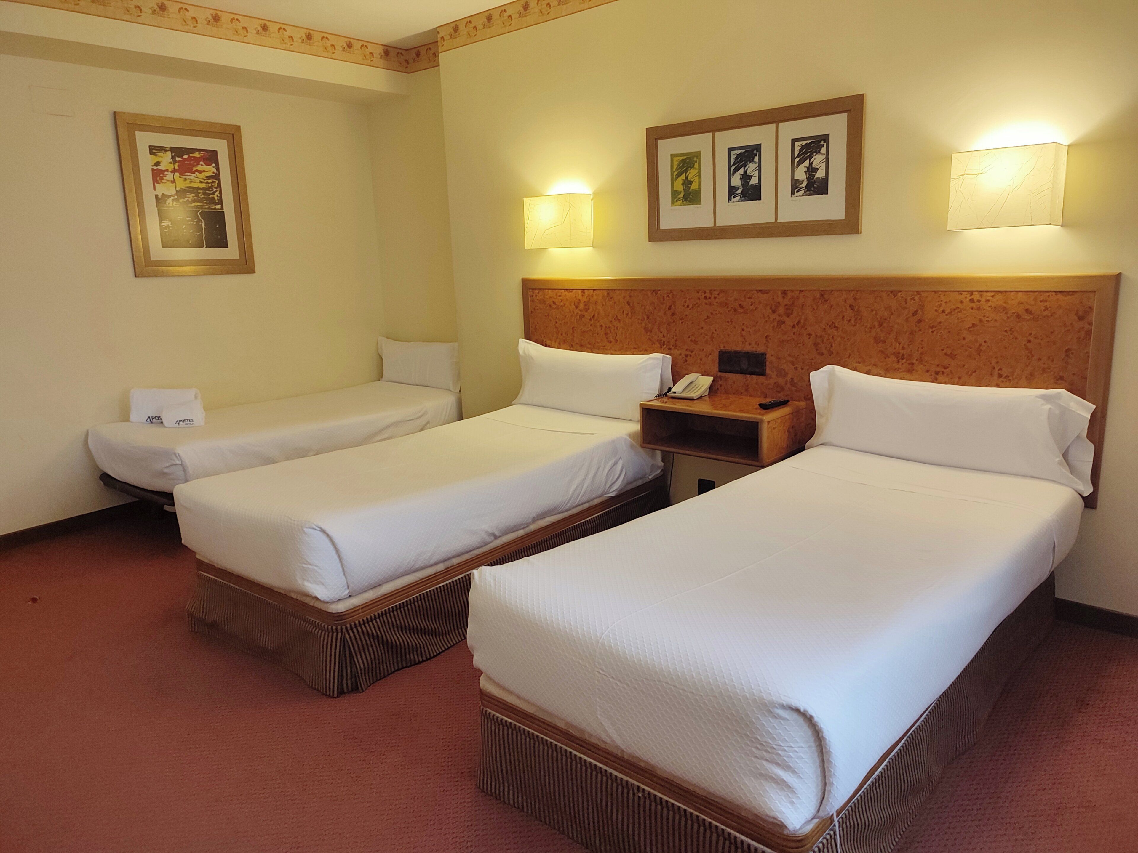 Une chambre d’hôtel avec deux lits simples, une tête de lit en bois, une table de chevet avec un téléphone et une télécommande, et des lampes fixées au mur.