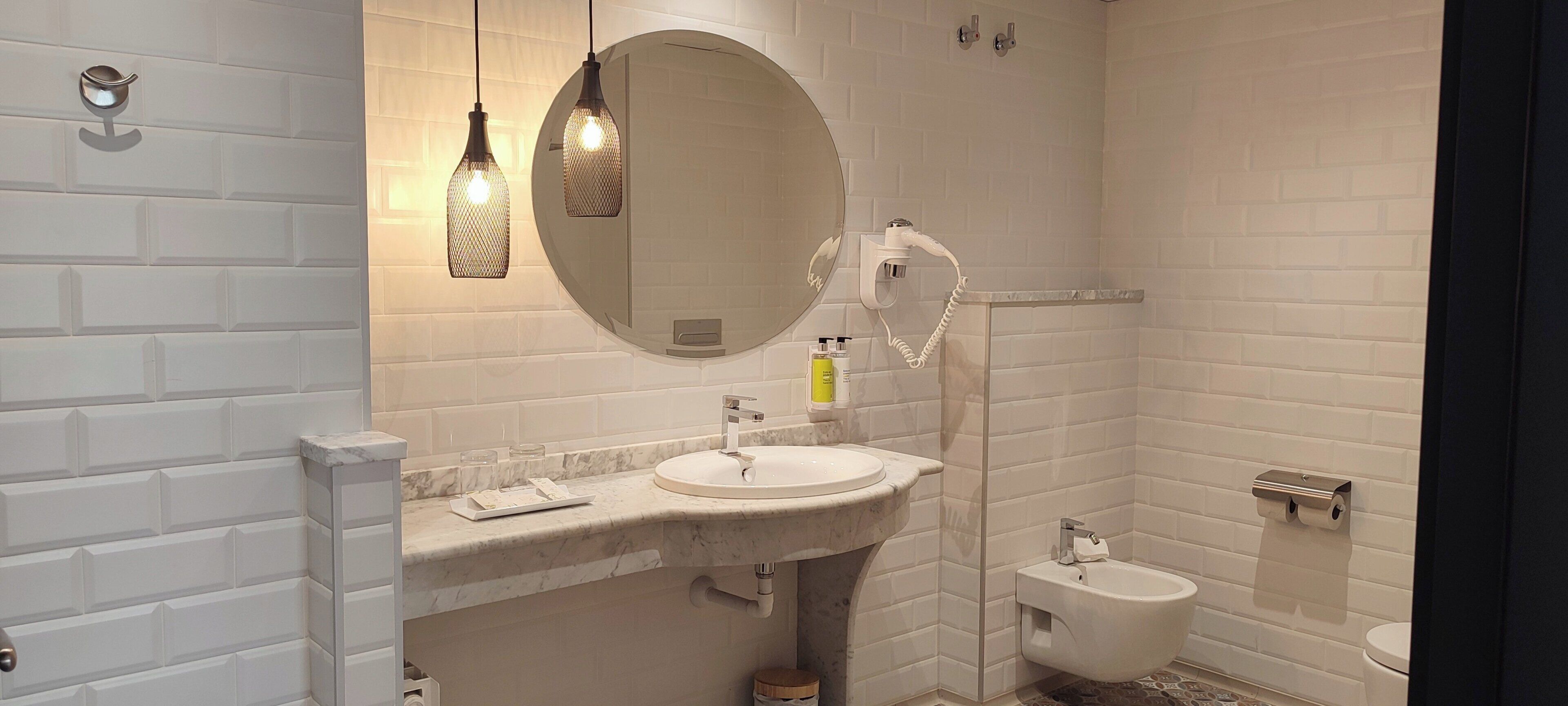 Un baño moderno con pared de azulejos tipo subway blancos, un lavamanos de mármol, un espejo redondo, dos luces colgantes, un cabezal de ducha de mano y un bidé.