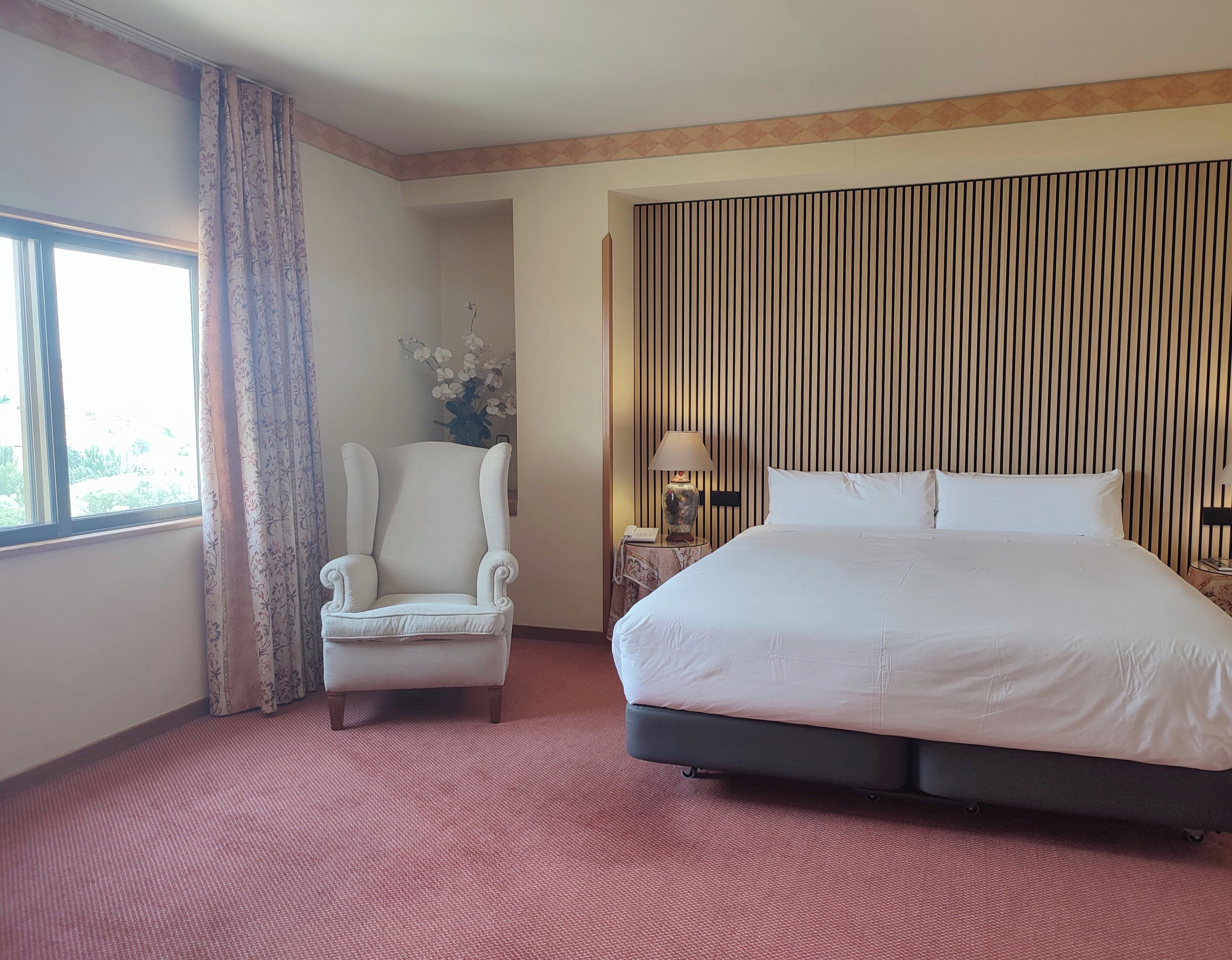 Habitación de hotel con una cama grande, una silla, una mesita de noche con lámpara y una ventana con cortinas.