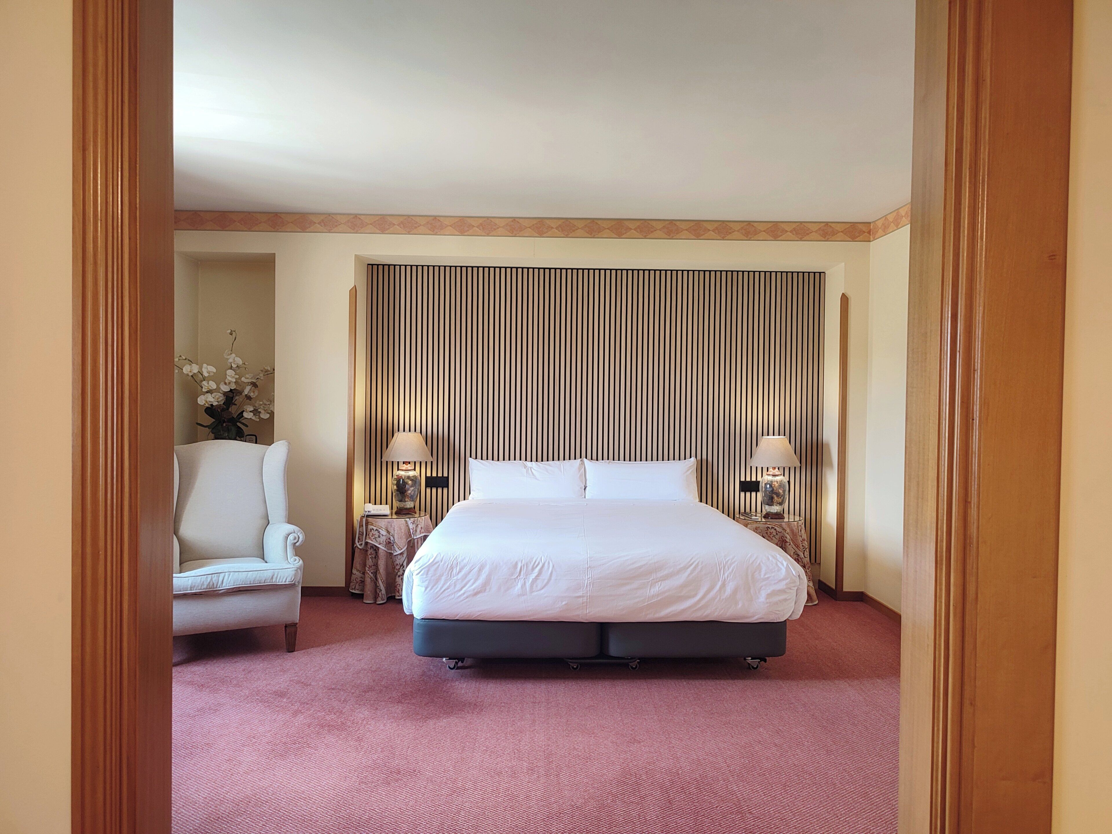 A hotel room with a large bed, two bedside tables with lamps, a chair, and a striped headboard.