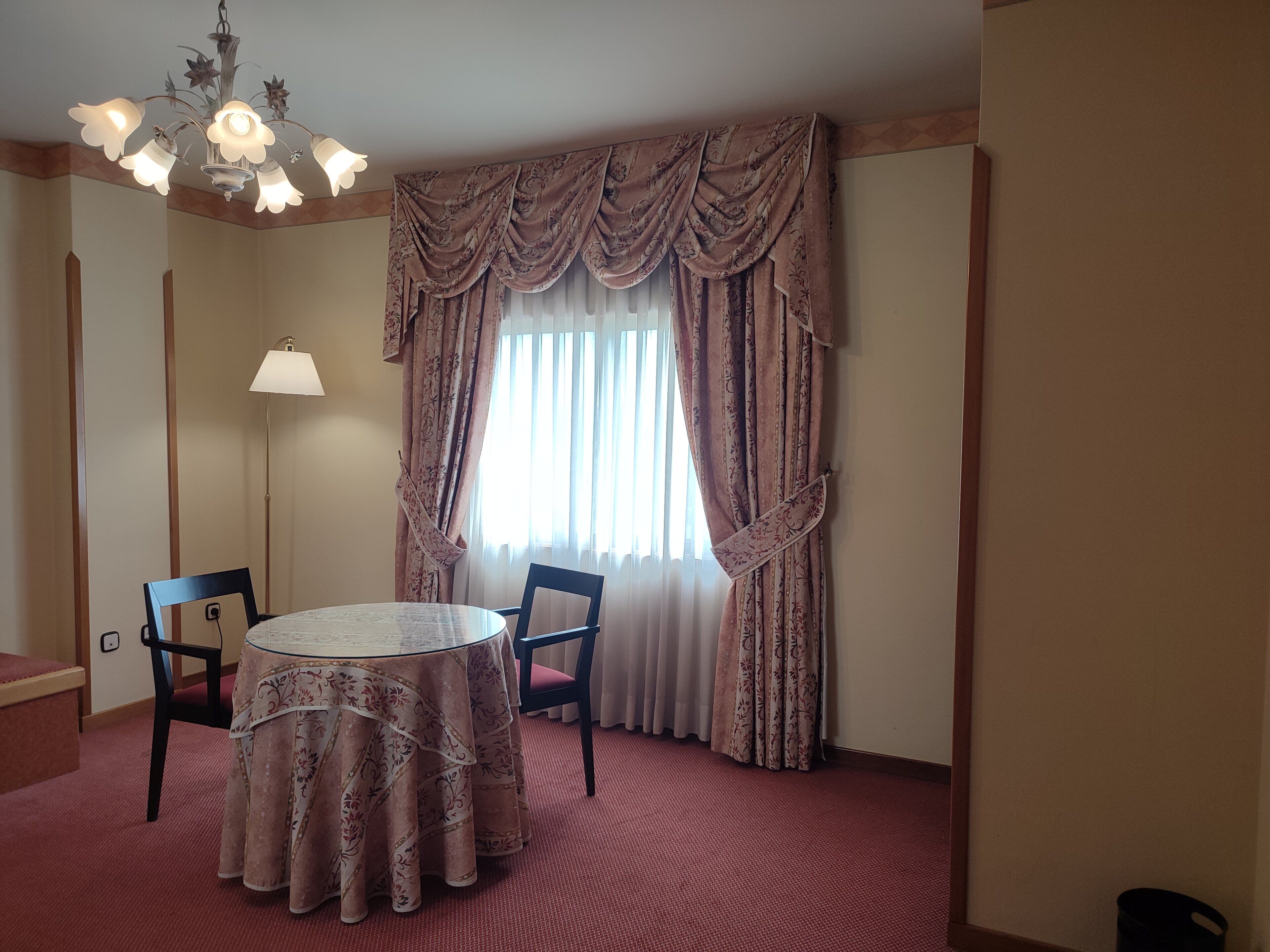 Une chambre d’hôtel avec une table ronde, deux chaises, un lustre et une grande fenêtre avec des rideaux.