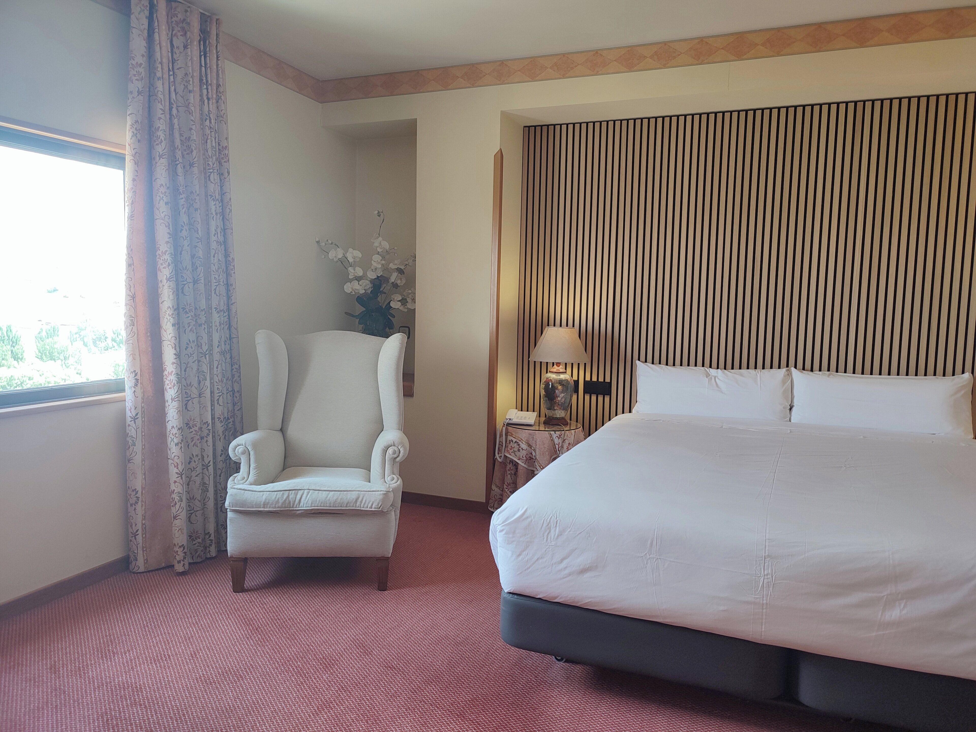 Habitación de hotel con una cama grande, una silla, una mesita de noche con lámpara y una ventana con cortinas.