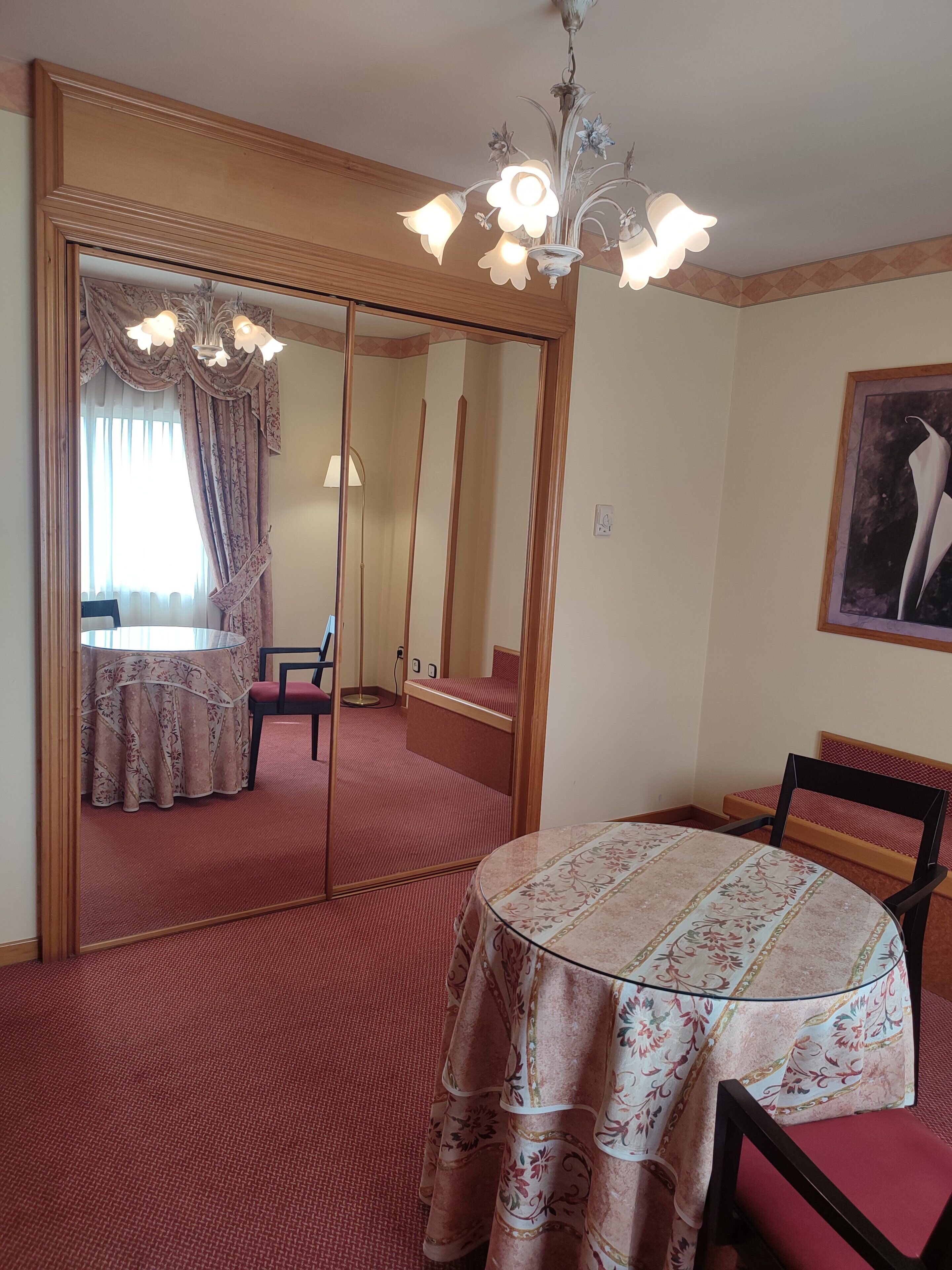 Una habitación con una mesa redonda, alfombra roja, un espejo grande y una araña.