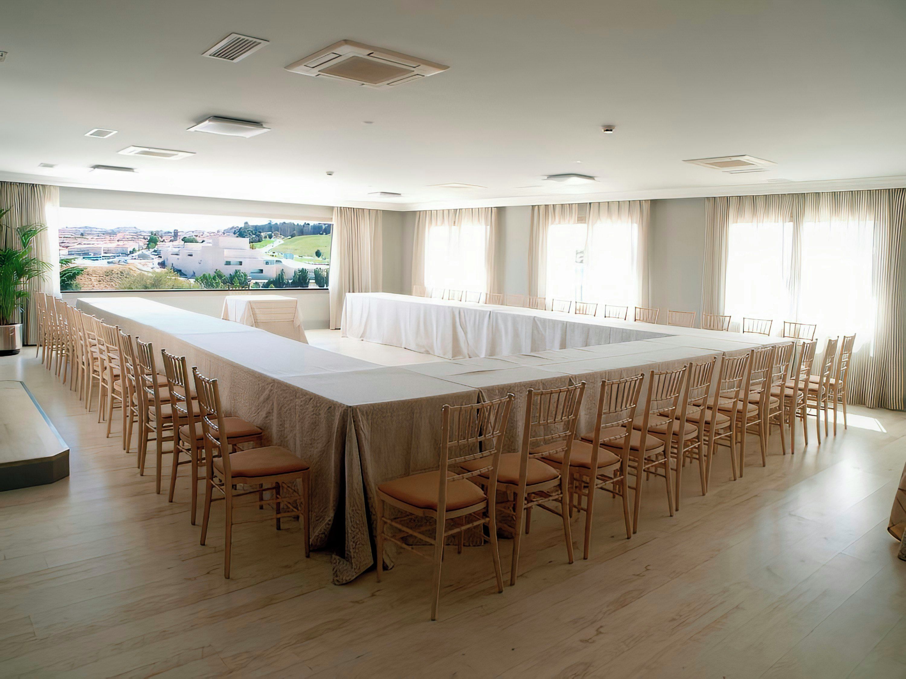 Un salón de conferencias con una larga mesa y sillas, ventanas amplias con cortinas y vistas a un paisaje.