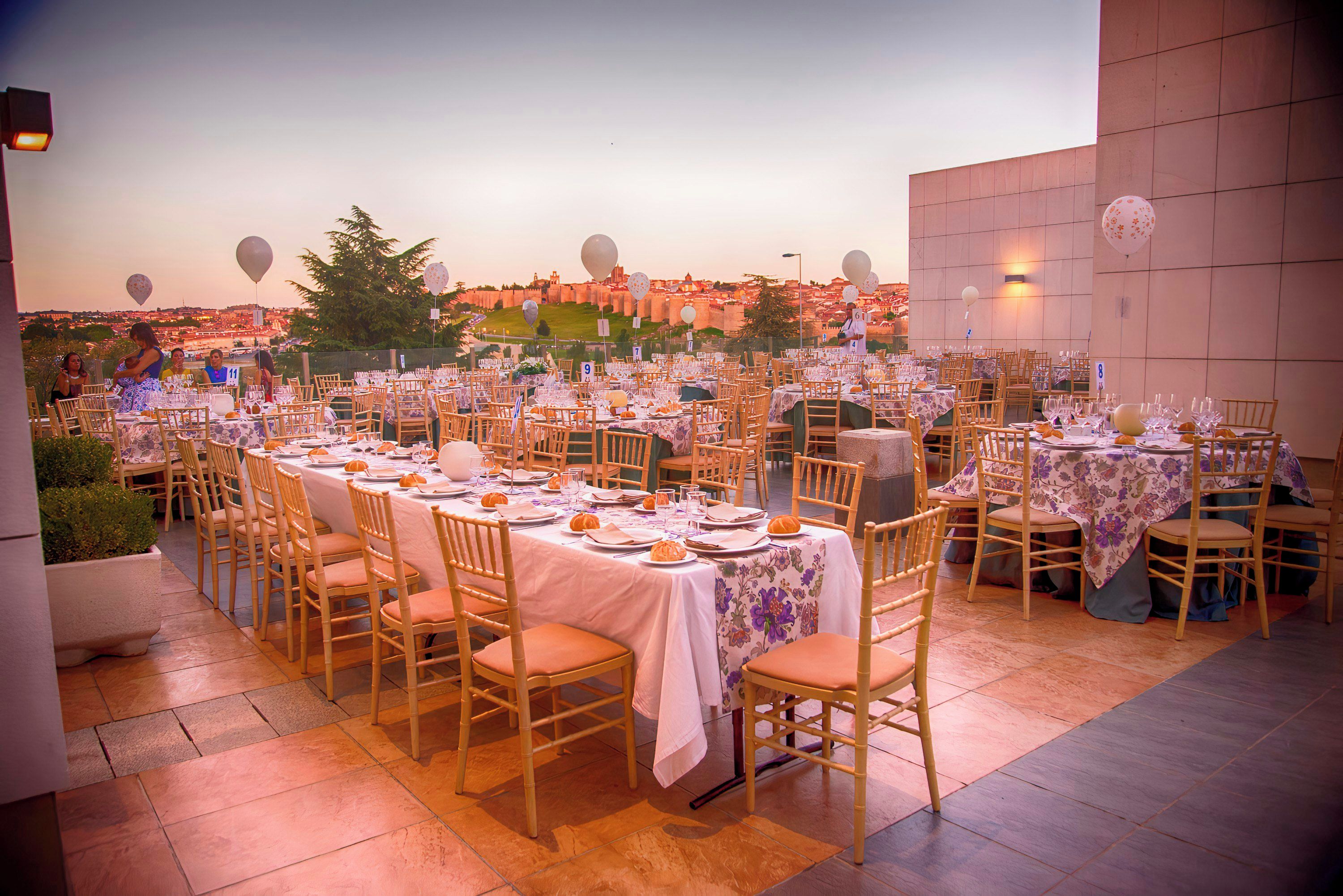 An outdoor dining area with multiple tables set for guests, chairs arranged around each table, and a view of the cityscape in the background.