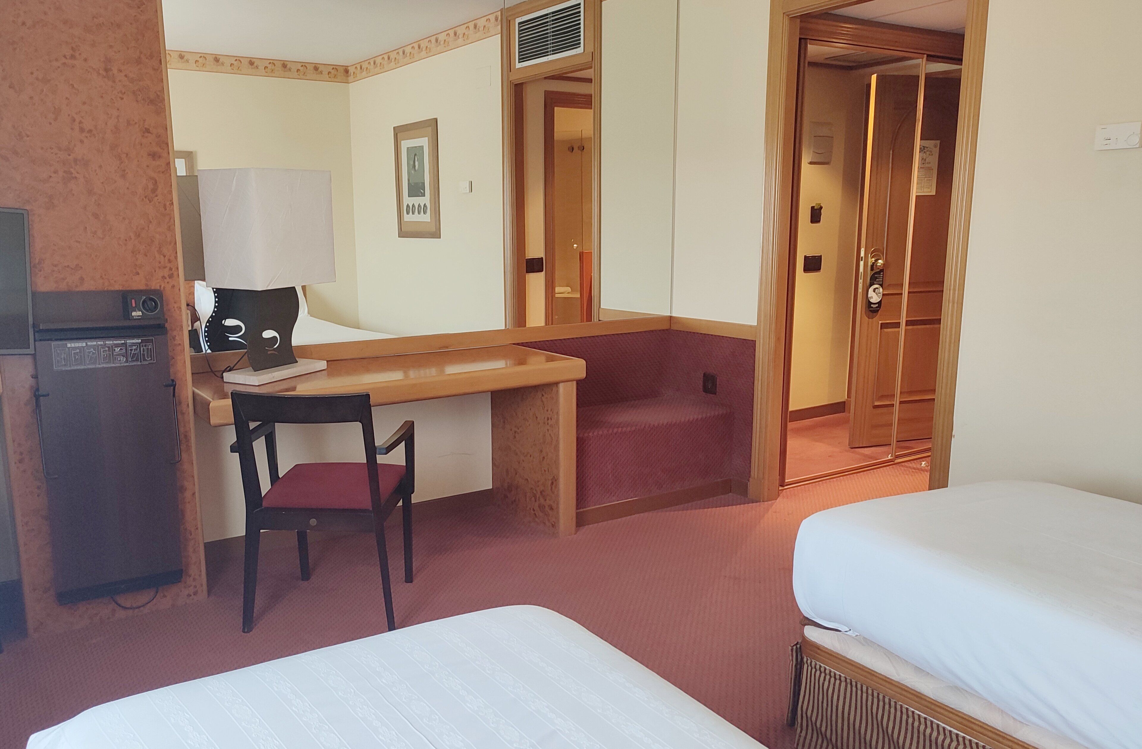 Habitación de hotel con cama, escritorio con silla, refrigerador pequeño y puerta que da a otra habitación.