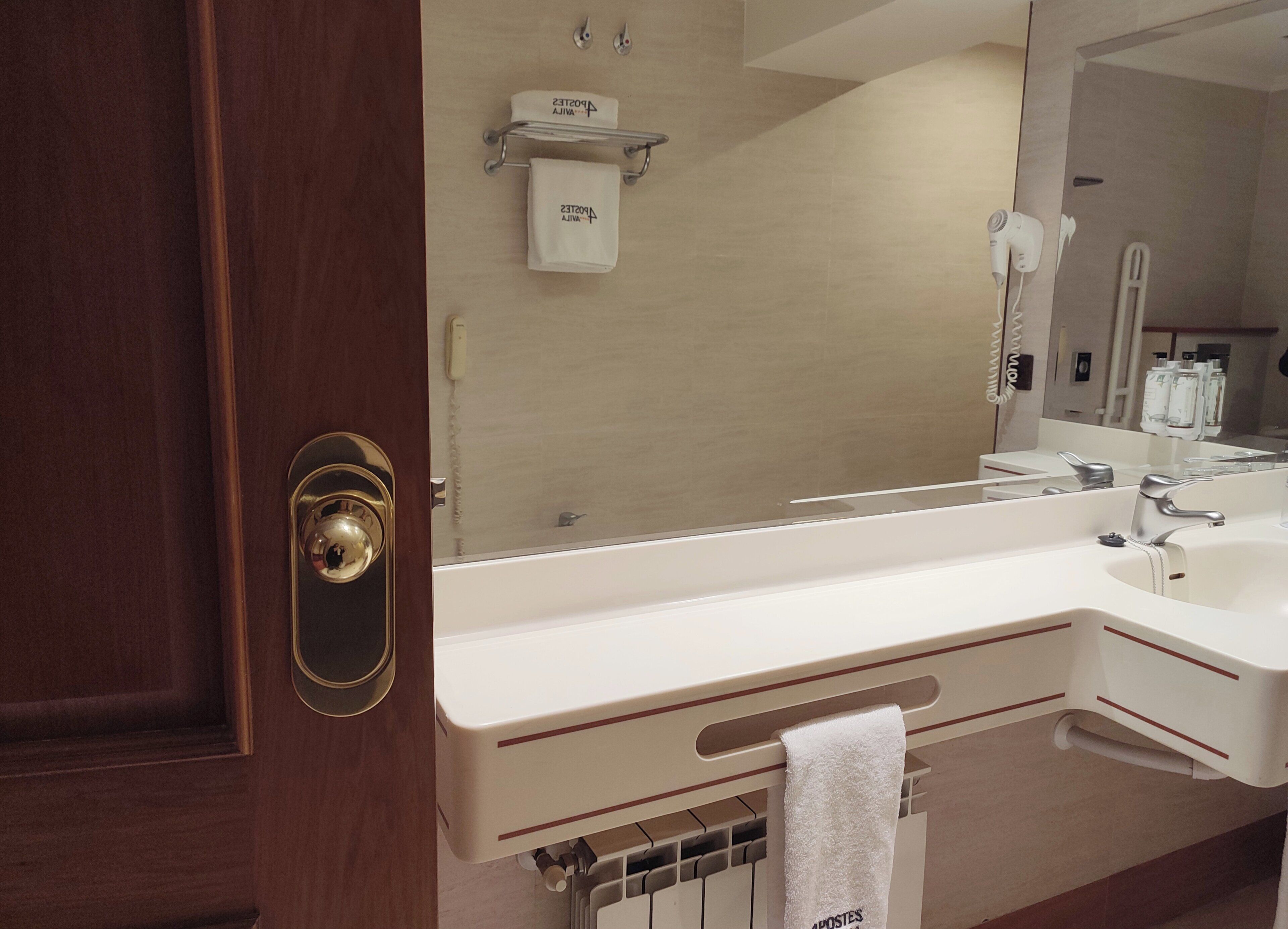 A hotel bathroom with a large mirror, a white sink, a hairdryer, and a towel rack.