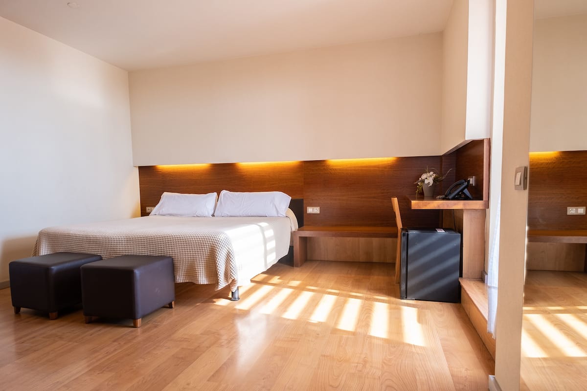 A modern hotel room with a bed, two ottomans, a desk, and a refrigerator.