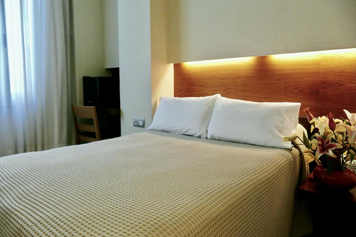 A hotel room with a neatly made bed, a wooden headboard, a bedside table with a vase of flowers, and a chair.