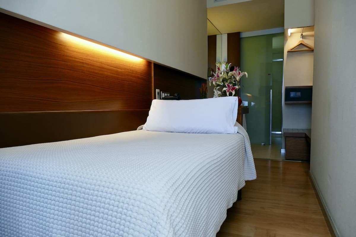 A hotel room with a large bed, a wooden headboard, a bedside table with a vase of flowers, and a microwave in the corner.