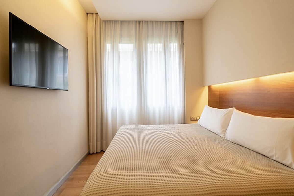 A hotel room with a large bed, a flat-screen TV mounted on the wall, and a window with sheer curtains.