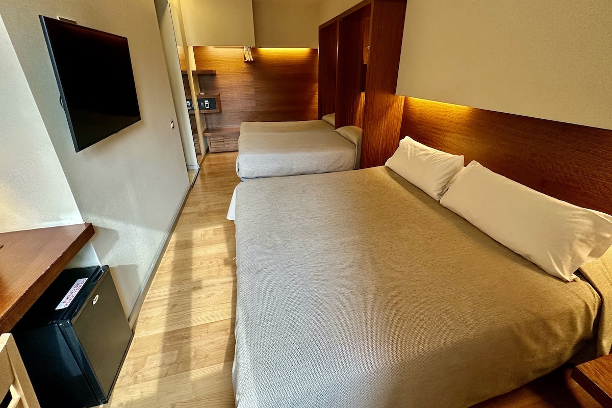 A hotel room with two beds, a small desk, a TV, and a wooden wardrobe.