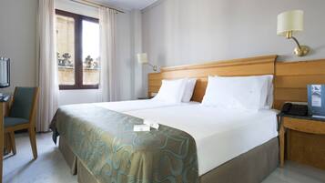 Habitación de hotel con una cama grande, un escritorio con silla, una ventana con cortinas y dos lámparas de pared.