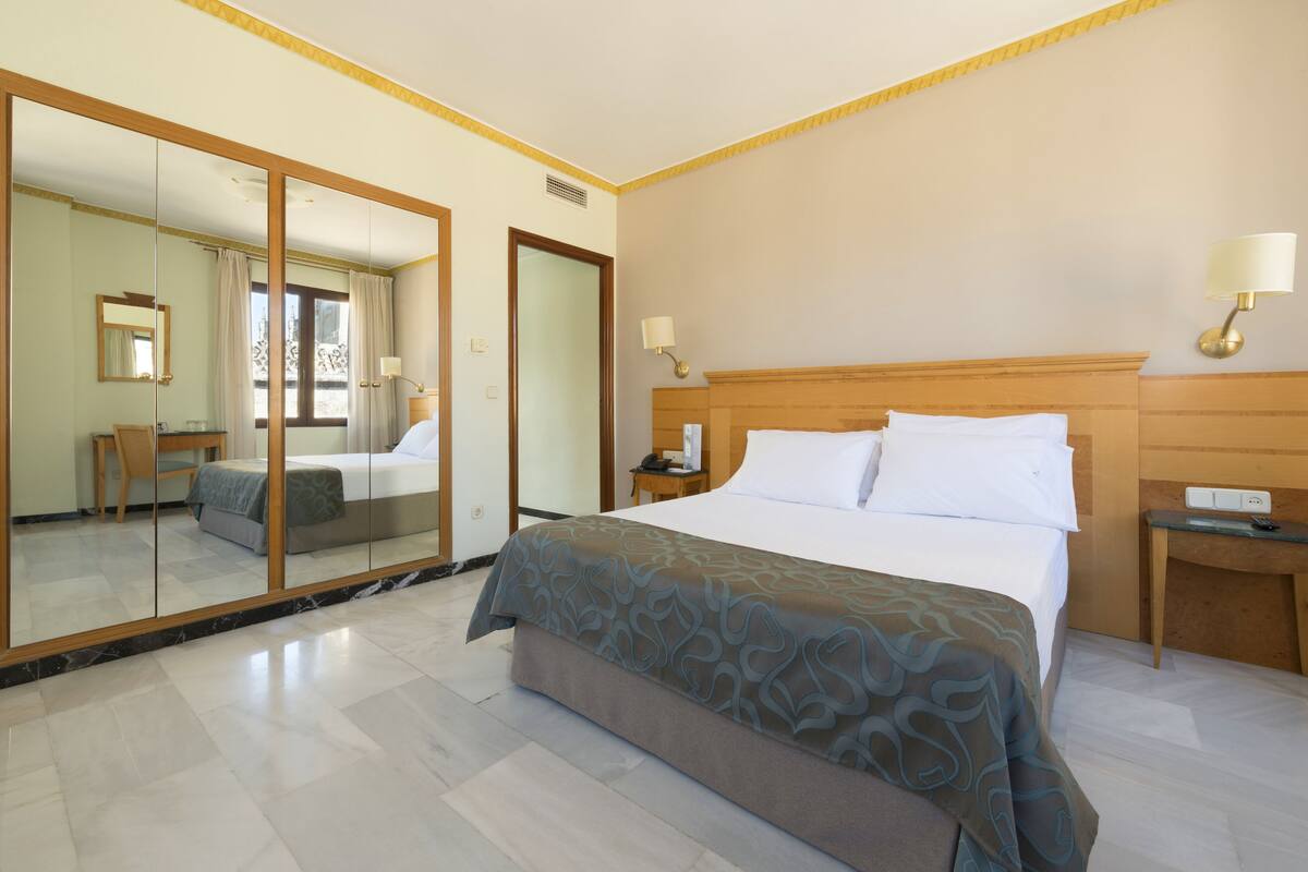 Une chambre d’hôtel avec un grand miroir, un lit avec du linge de maison blanc, une table de chevet avec une lampe et une tête de lit en bois.