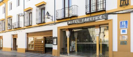 A hotel facade with a sign reading "HOTEL MAMONIDES," featuring balconies, a large glass entrance, and a cobblestone street.