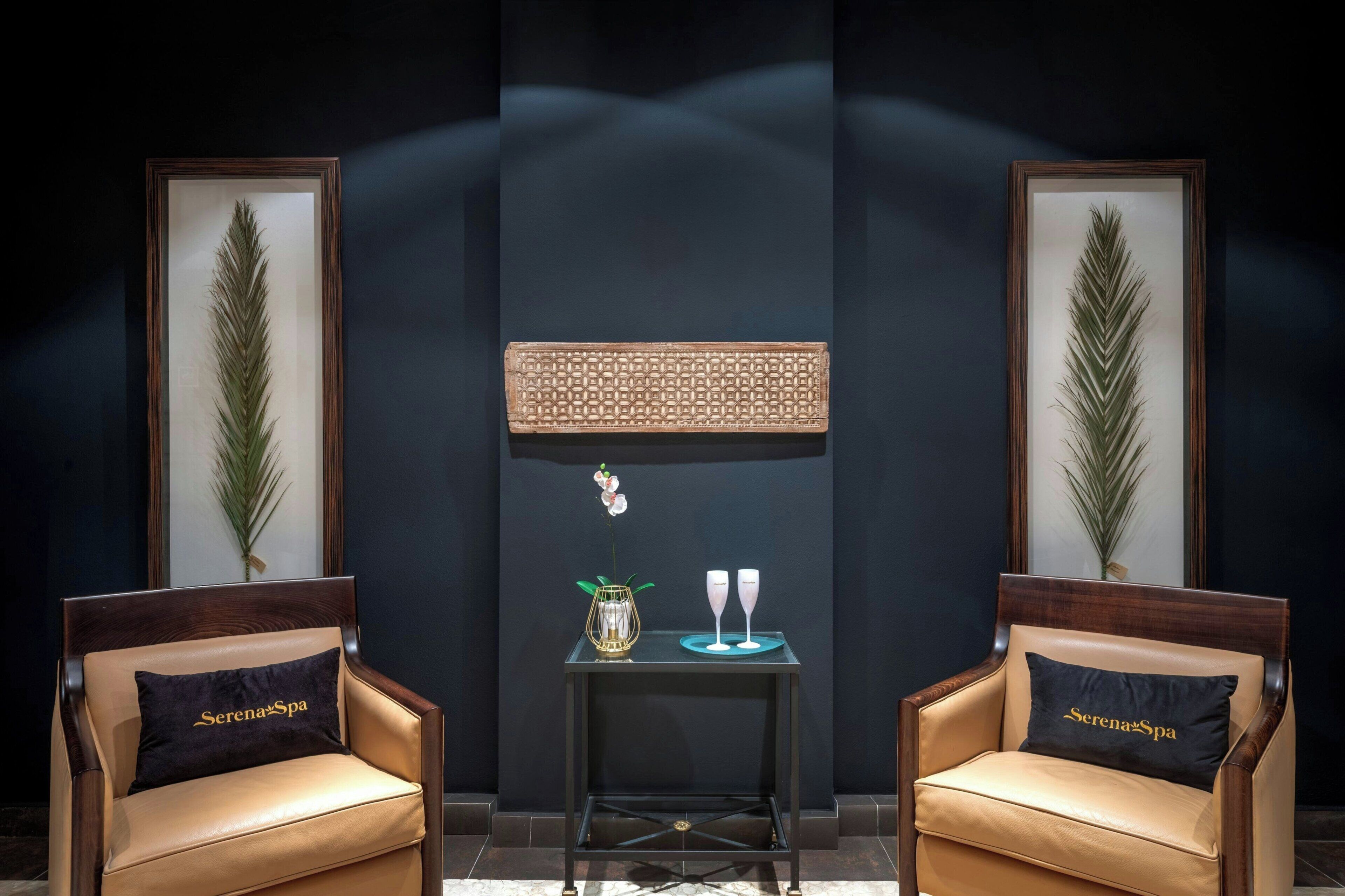 A room with two leather chairs, a small table with a vase and two glasses, framed artwork on the walls, and a dark floor.