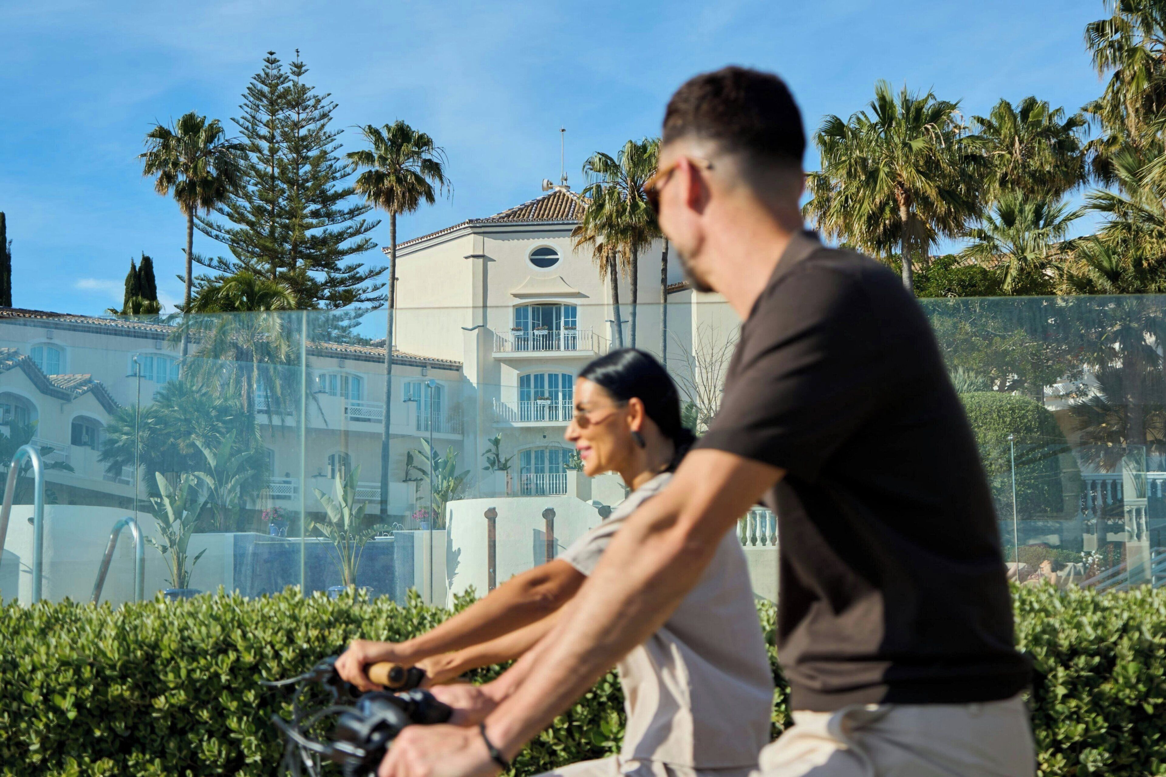 Dos personas pedaleando una bicicleta estática, con un edificio y palmeras al fondo.