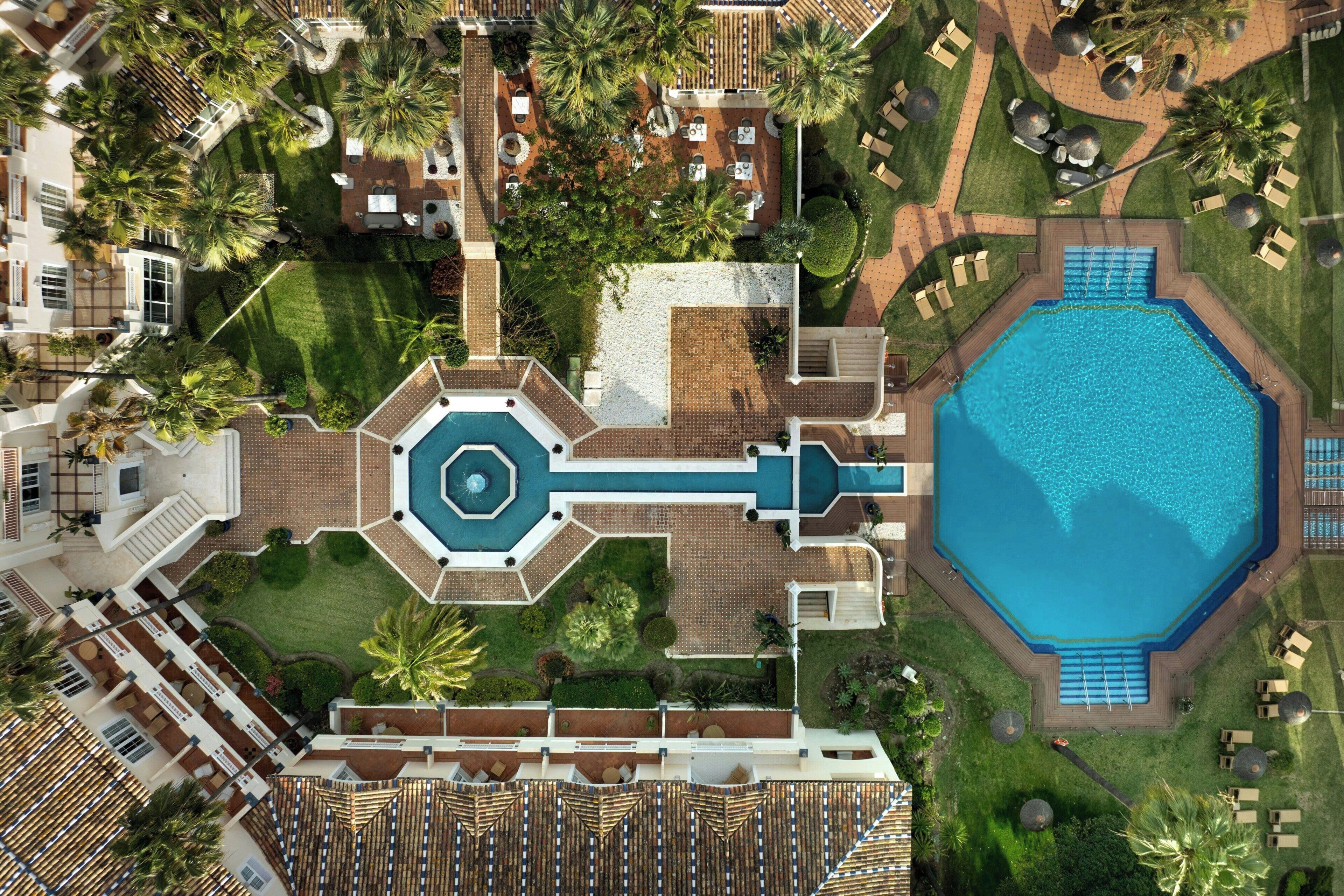 An aerial view of a resort with a central pool, multiple guest rooms, and palm trees.