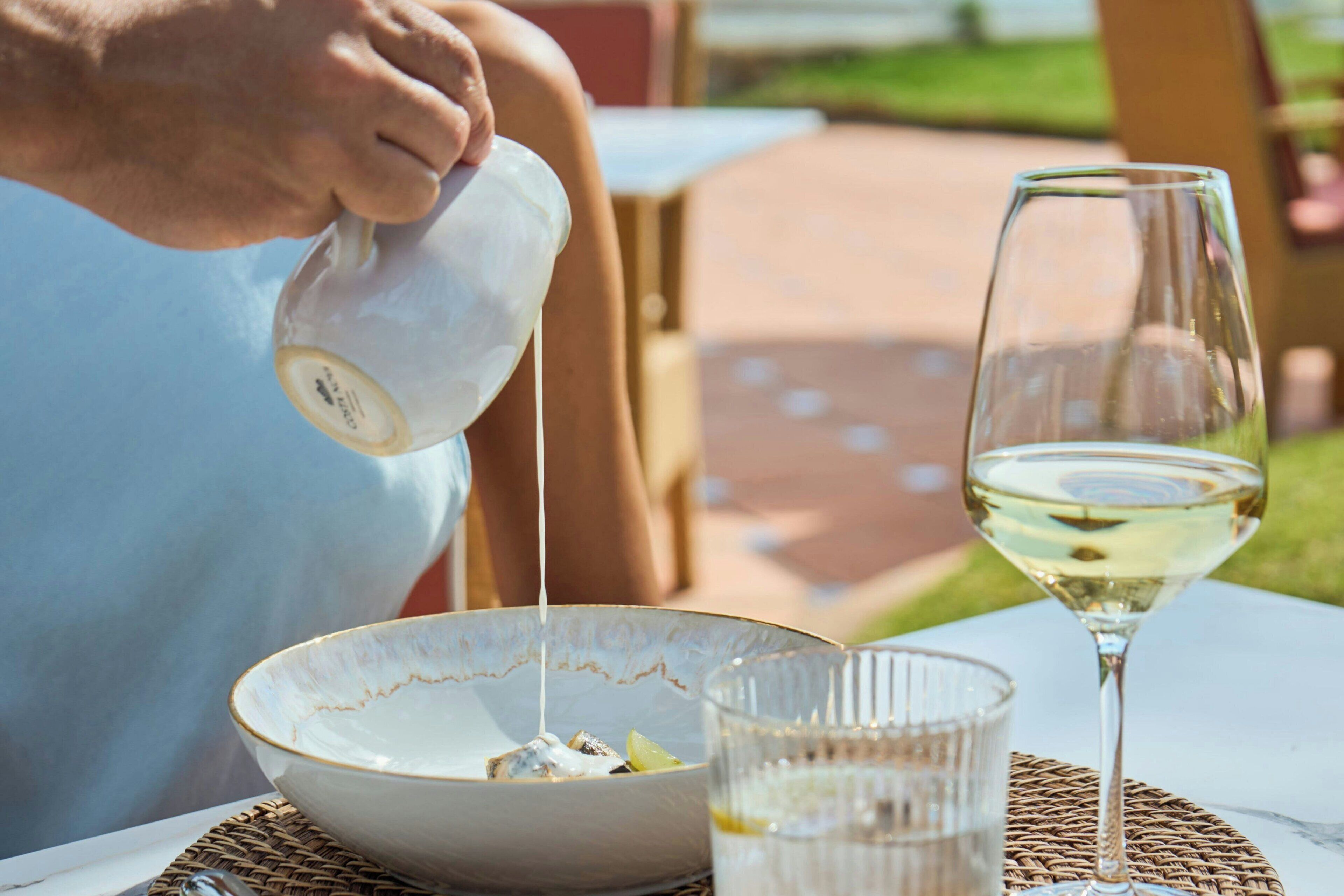 A person pouring a clear liquid into a bowl, a glass of white wine, and a glass of water on a table.
