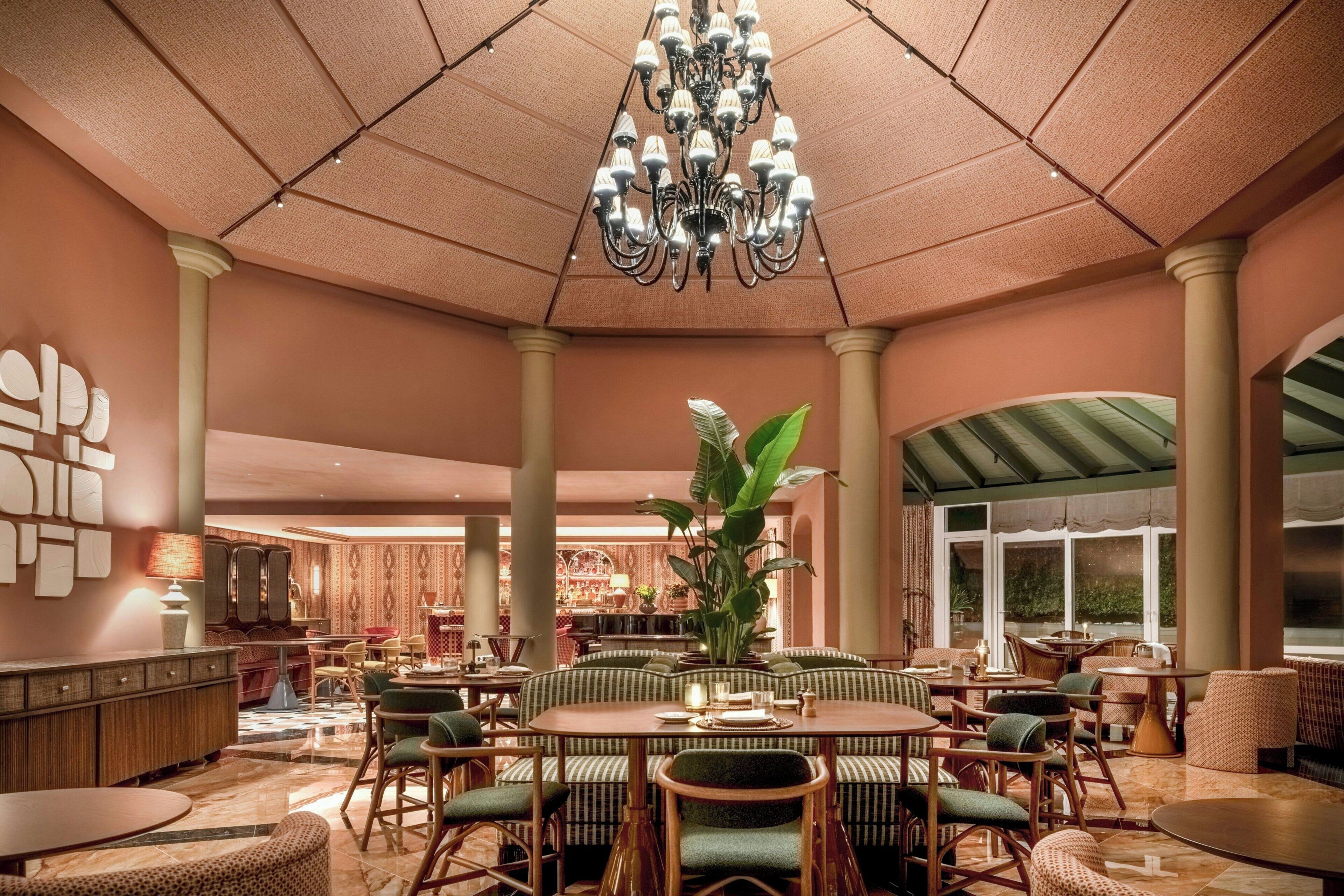 A spacious indoor area with a central round table surrounded by chairs, a large chandelier, and a plant.