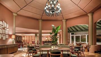 A spacious indoor area with a central round table surrounded by chairs, a large chandelier, and a plant.