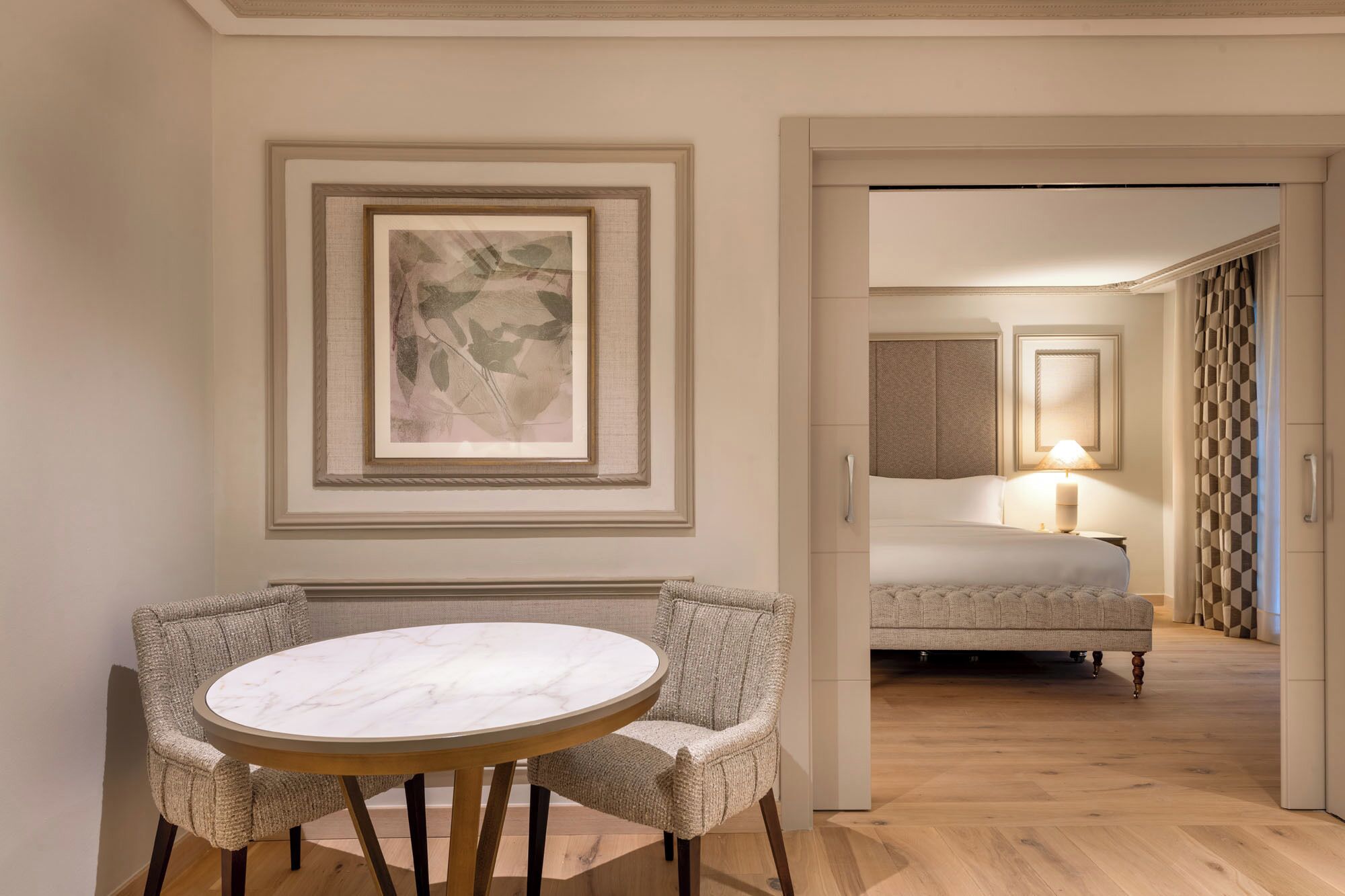Una habitación de hotel con una cama, una mesita de noche con lámpara, una obra de arte abstracto enmarcada en la pared, y una mesa redonda con sillas al frente.
