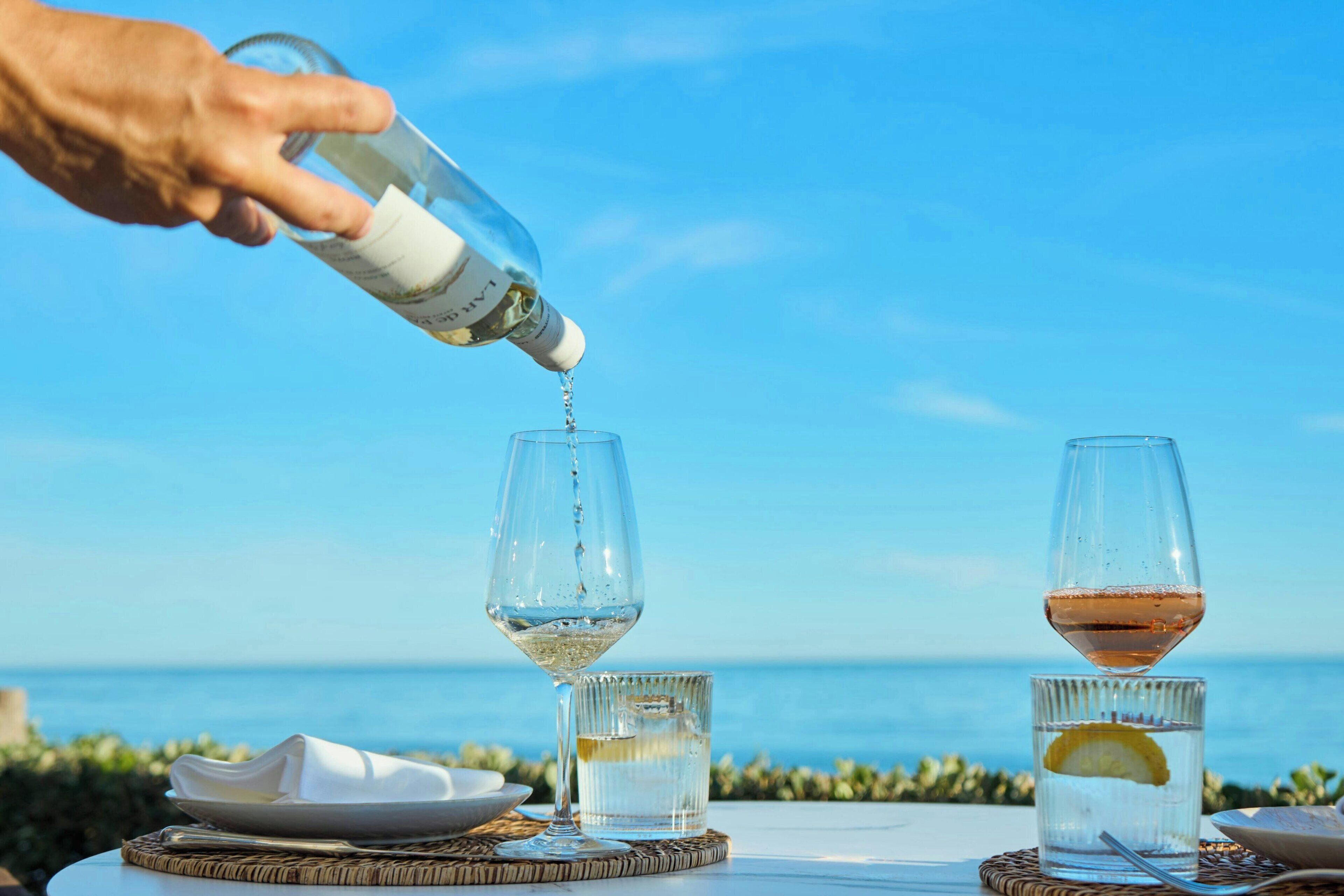 A hand pouring a clear liquid into a glass, with a view of the sea in the background.