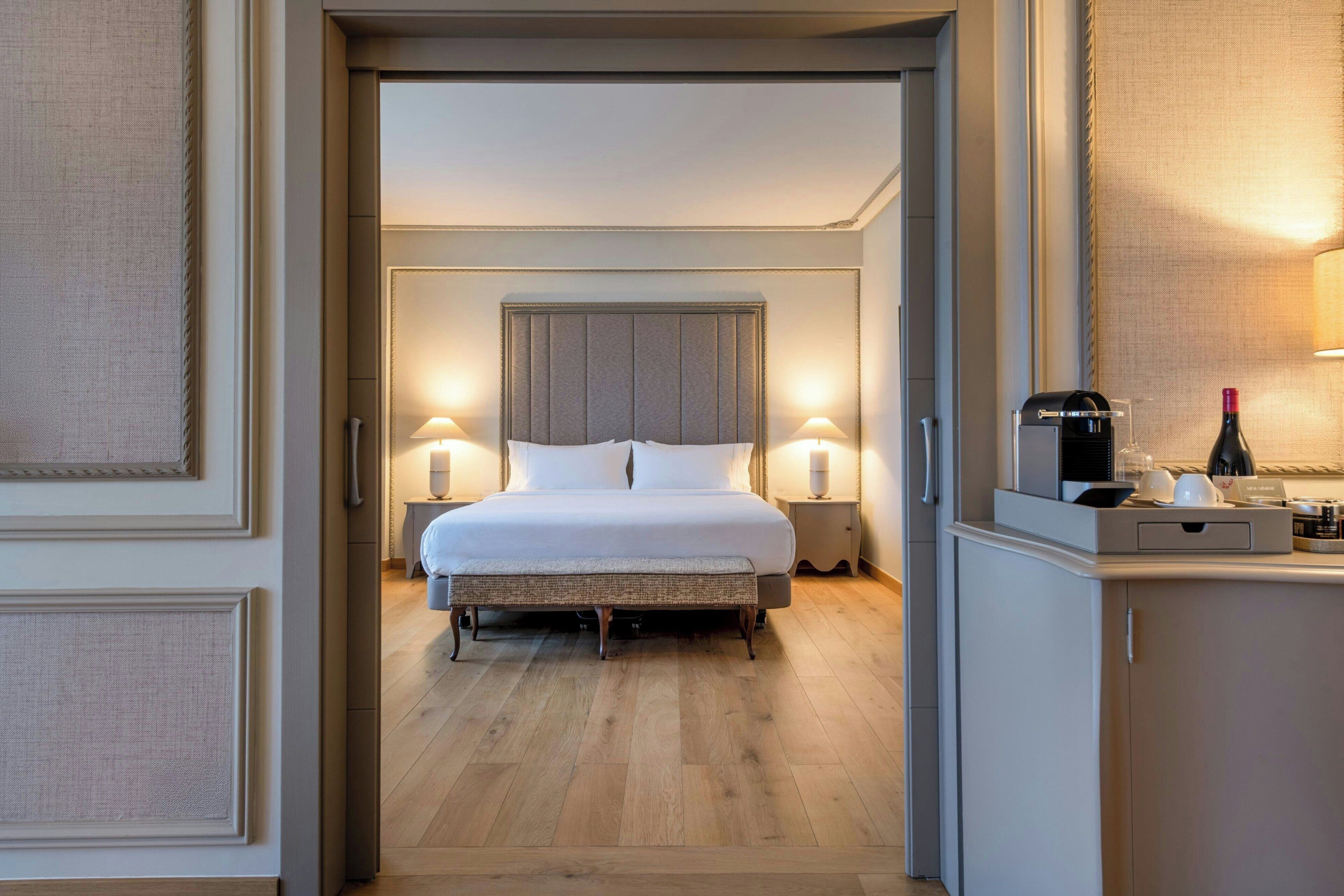 Habitación de hotel con cama, lámparas de noche, mesita de noche, cafetera y una botella de vino.