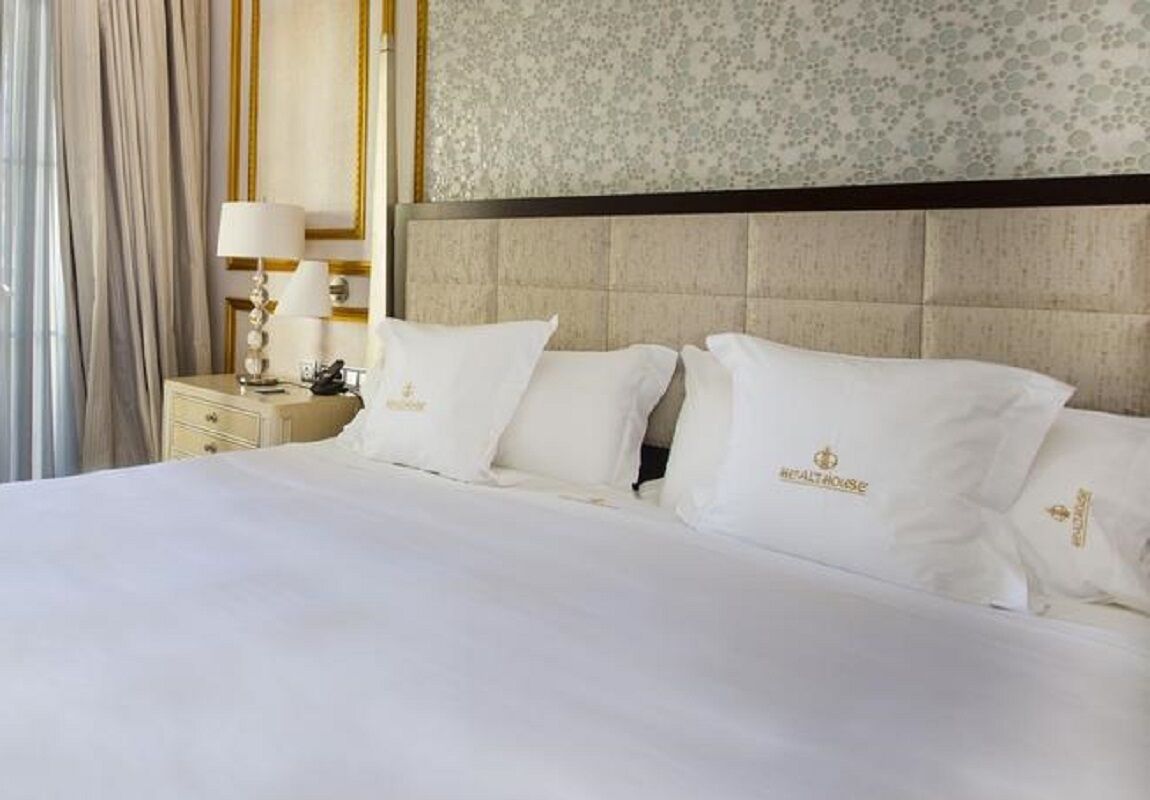 A hotel room with a large bed, two white pillows with a logo, a nightstand with a lamp, and a floral-patterned wall.