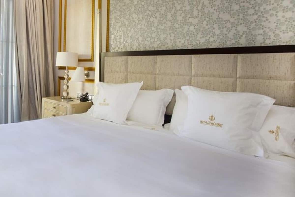 A hotel room with a large bed, two white pillows with a logo, a nightstand with a lamp, and a floral-patterned wall.