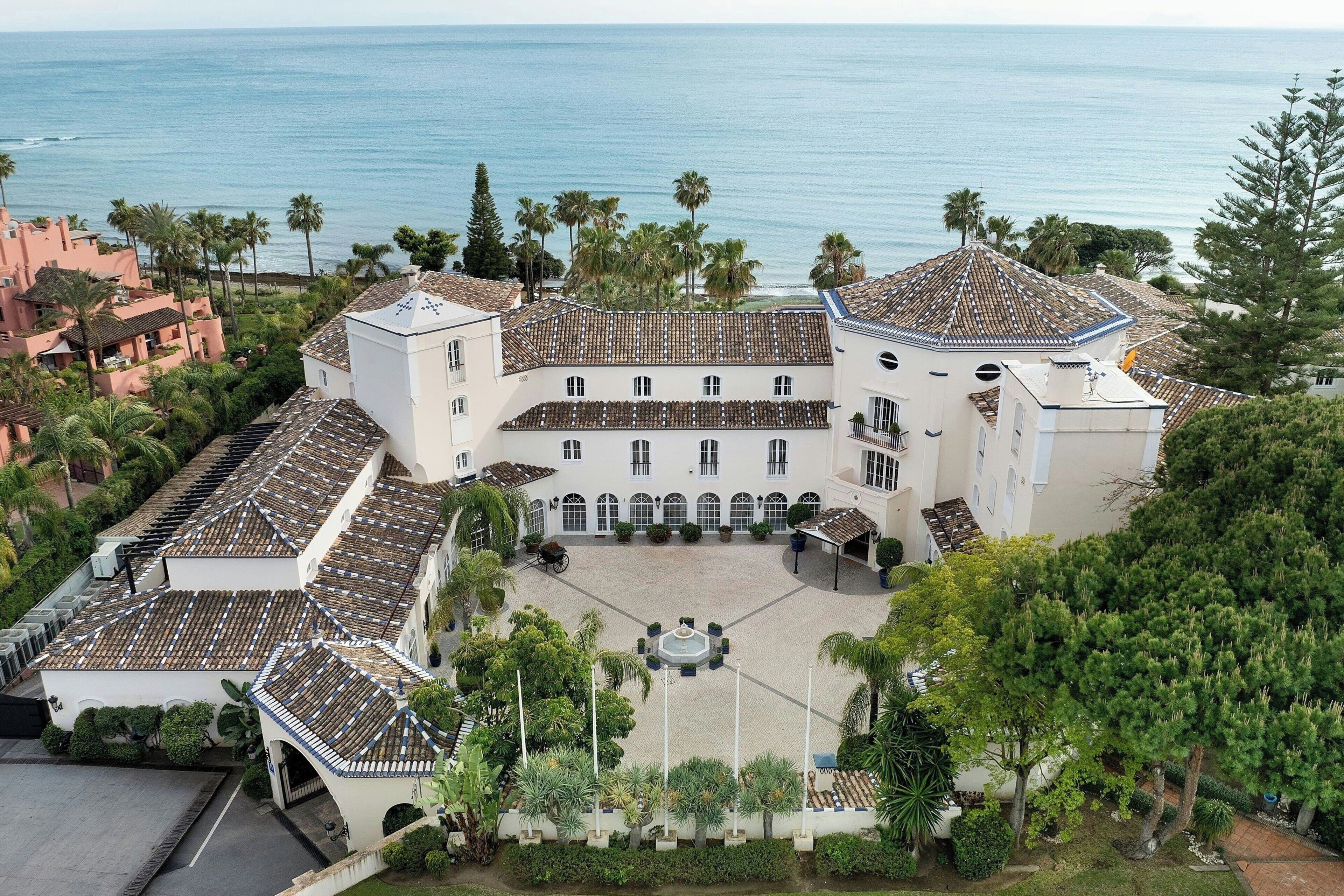 Un grand bâtiment blanc doté d’une cour intérieure, entouré de palmiers et offrant une vue sur l’océan.