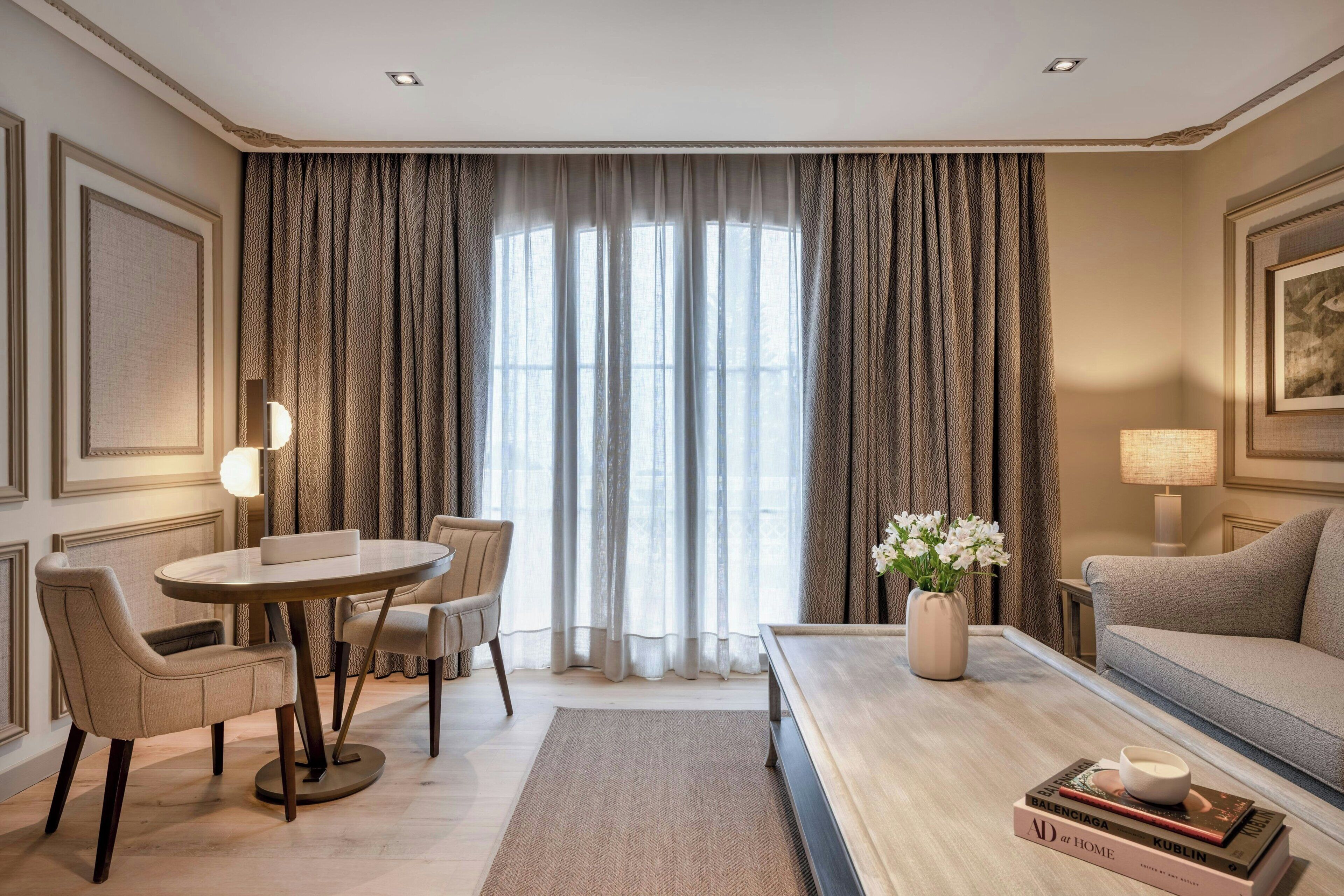Ein modernes Hotelzimmer mit einem runden Esstisch, zwei Stühlen, einer Couch, einem Couchtisch mit Büchern und einer Vase sowie einem großen Fenster mit Vorhängen.
