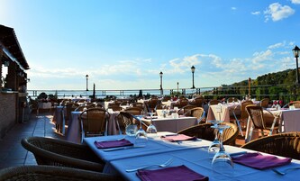 An outdoor dining area with tables set for two, wicker chairs, and a view of the sea.