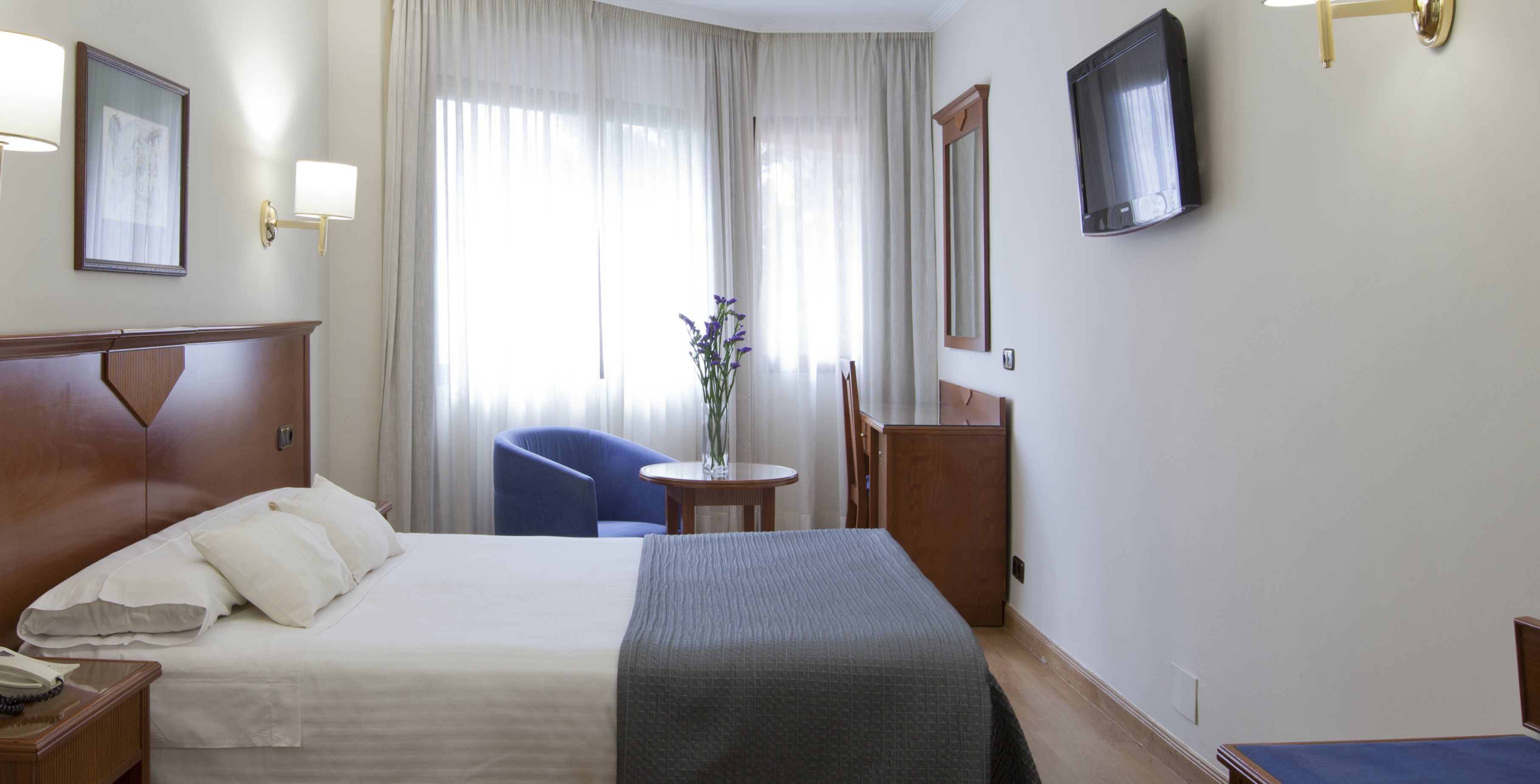 Hotelkamer met een bed, nachtkastje, lamp, stoel en een tafeltje met een bloevas.