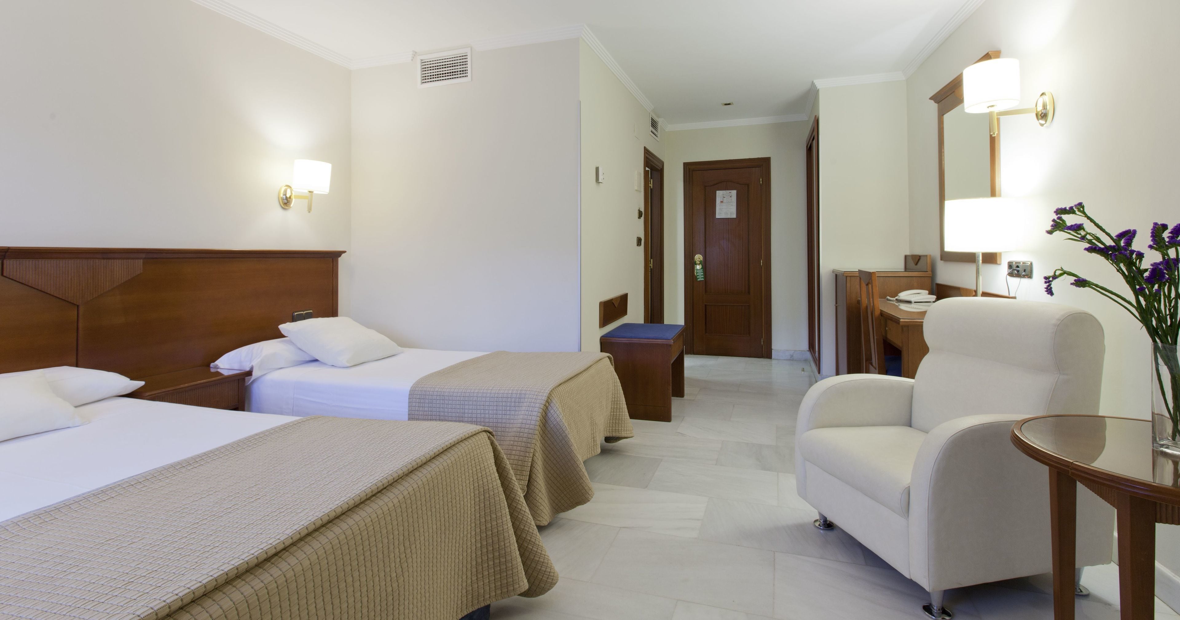 Habitación de hotel con dos camas, un espacio de descanso con una silla y una mesa, y un baño a simple vista al fondo.