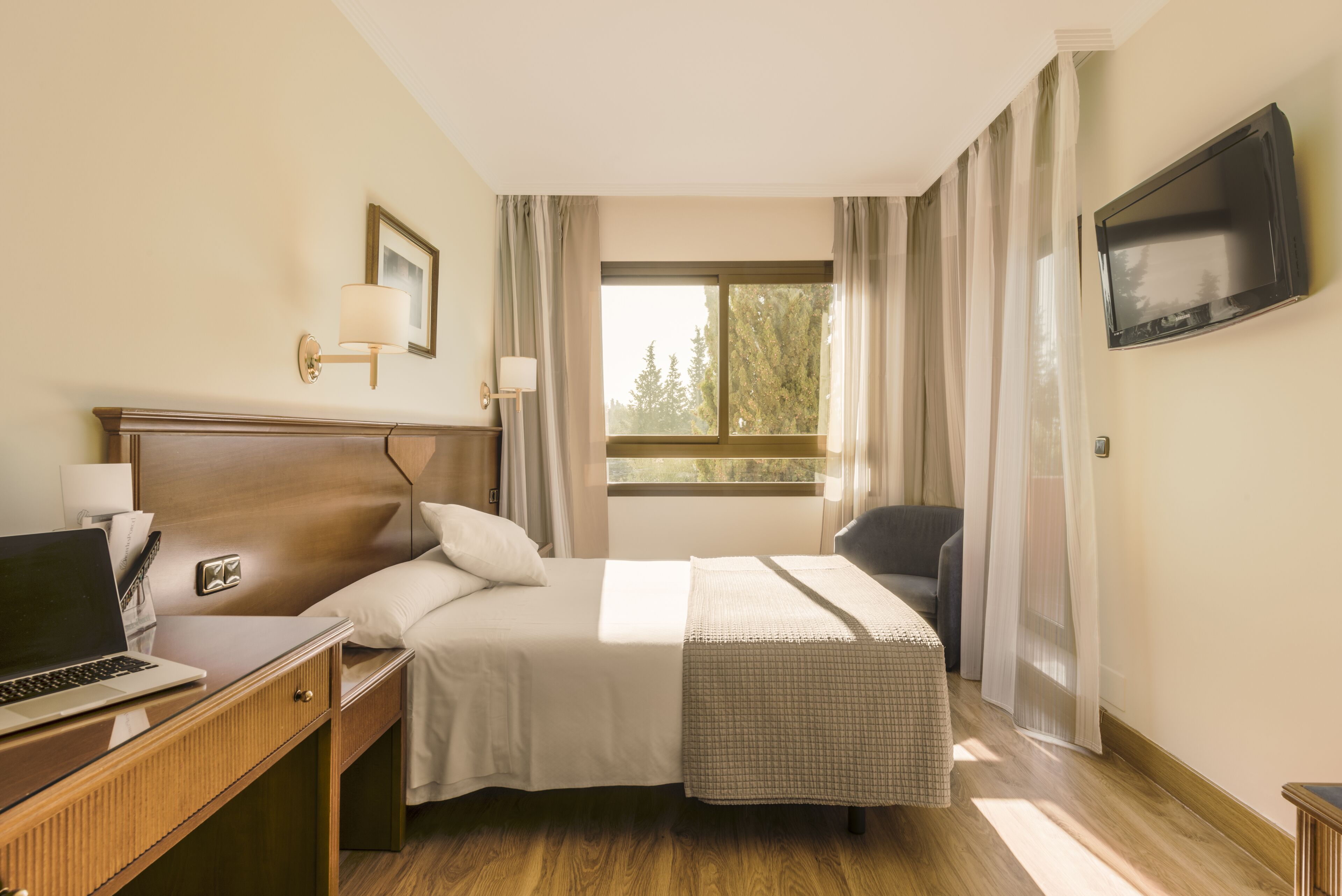 Habitación de hotel con cama, escritorio con computadora portátil, televisor montado en la pared y ventana con cortinas.