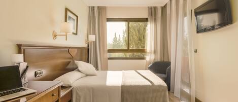 Habitación de hotel con cama, escritorio con computadora portátil, televisor montado en la pared y ventana con cortinas.