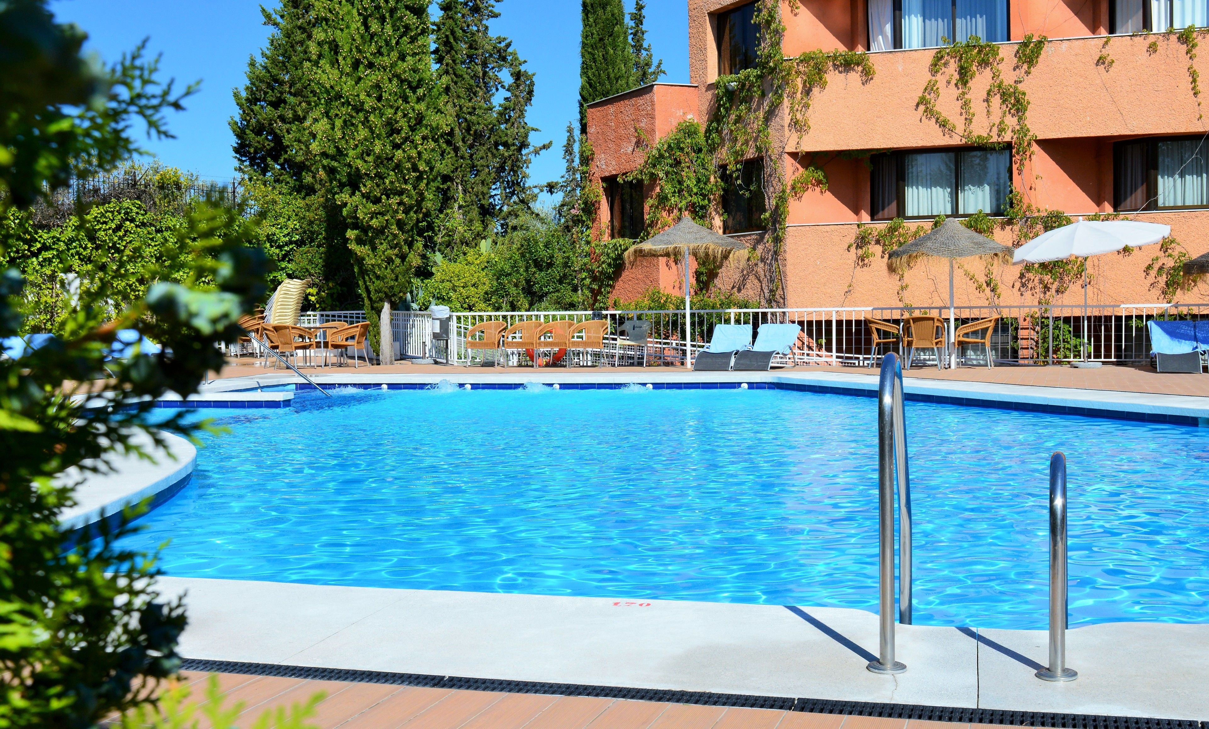 Une piscine entourée d’une clôture et d’arbres, avec des chaises longues et des parasols.