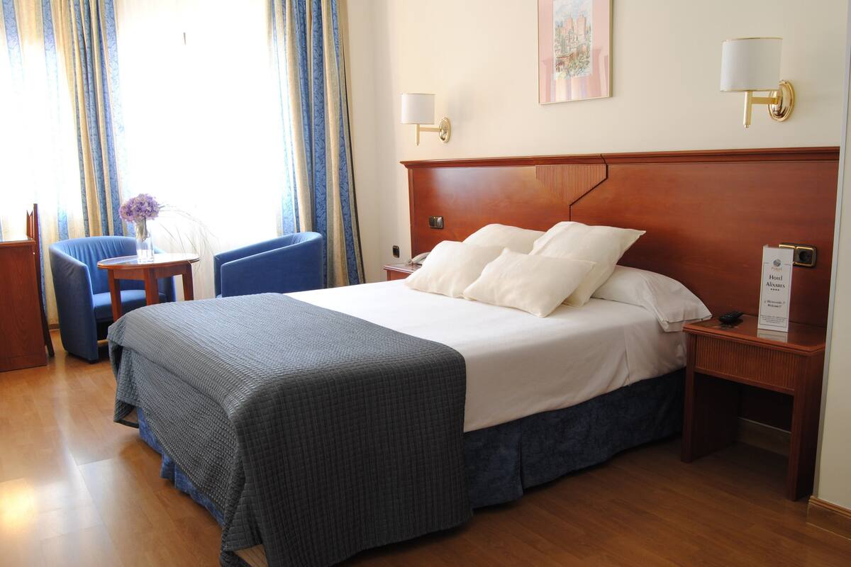 A hotel room with a large bed, a desk with a chair, a small table with a vase, and a nightstand with a lamp.