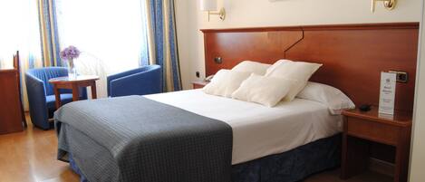 Habitación de hotel con una cama grande, un escritorio con una silla, una mesita con un florero y una mesita de noche con una lámpara.
