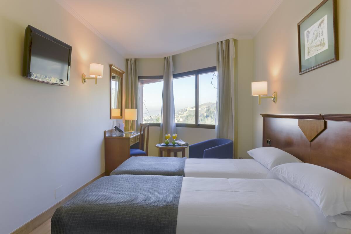 A hotel room with two beds, a desk, a chair, a TV, and a window with a city view.