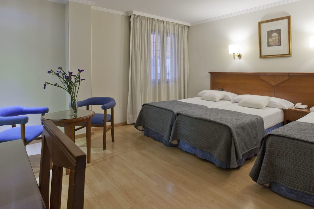 A hotel room with two beds, a desk with a chair, a vase with flowers, and a window with curtains.